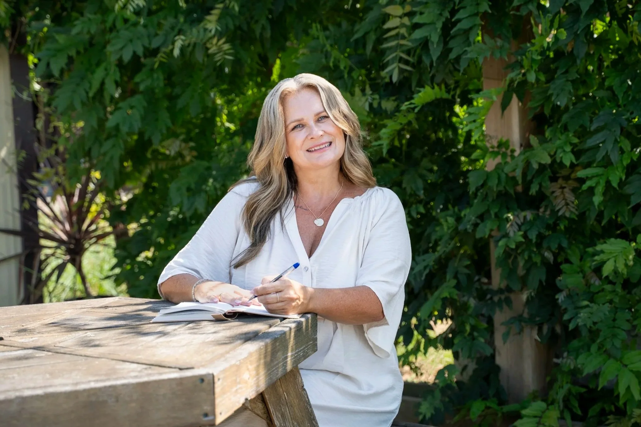 A woman with long wavy blonde hair wearing a white shirt sits outdoors at a wooden table with greenery in the background. She is smiling, holding a pen, and has an open notebook in front of her.