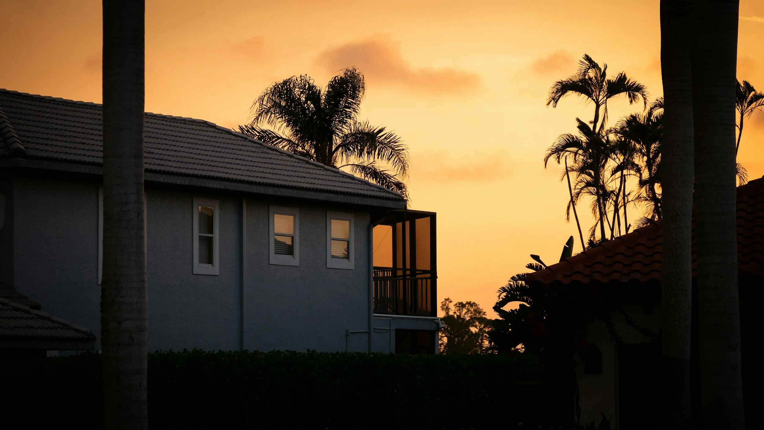 A residential house with a tiled roof and windows, surrounded by palm trees, during a sunset with an orange sky.