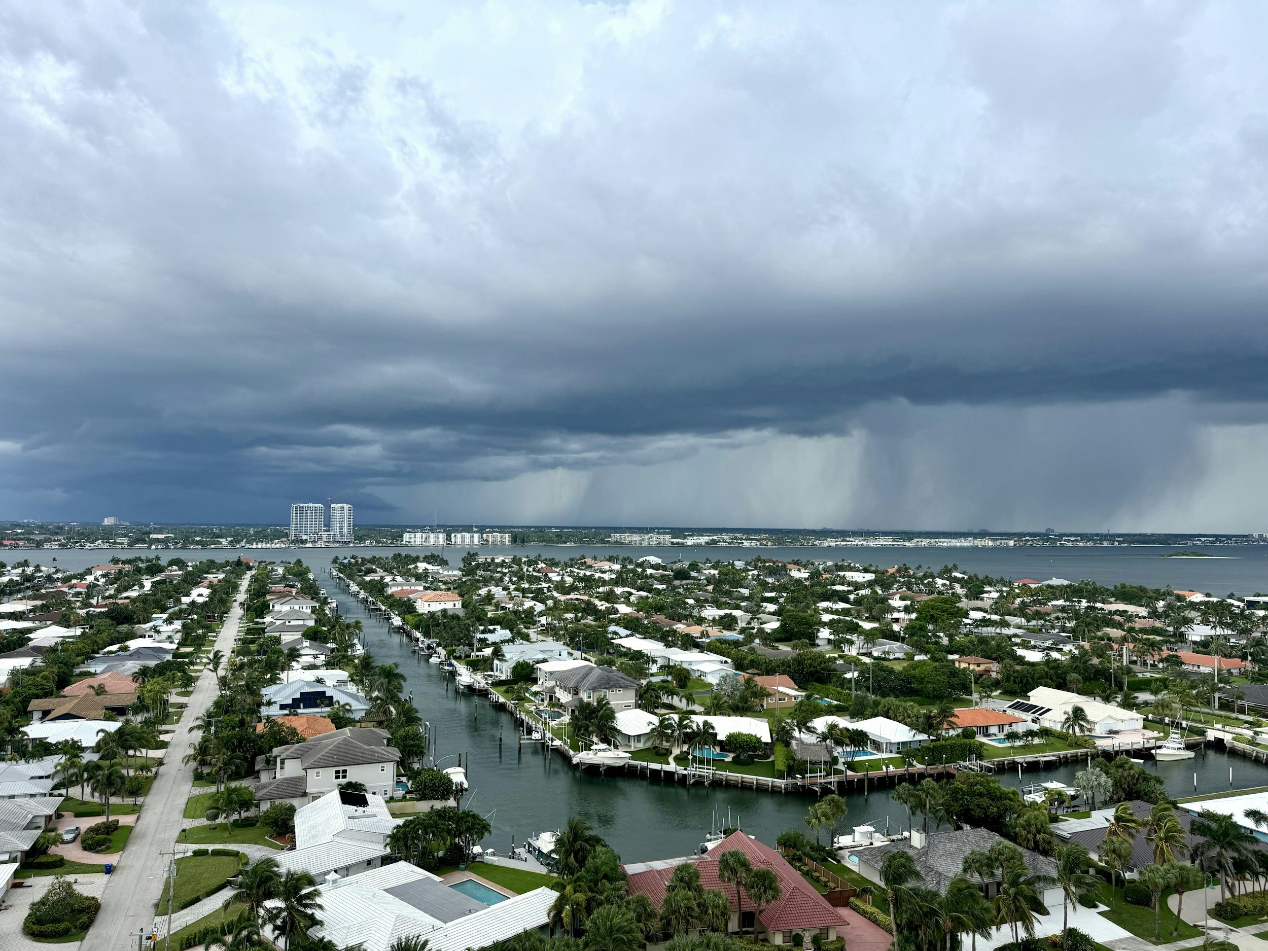 A residential neighborhood with boats docked along a canal, under a cloudy sky with dark storm clouds over a city skyline in the distance.