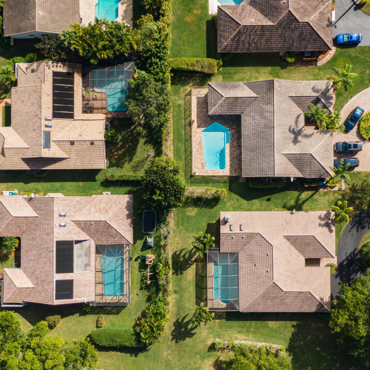Aerial view of several suburban houses with tiled roofs, some with swimming pools and screened-in patios, surrounded by green lawns and trees.