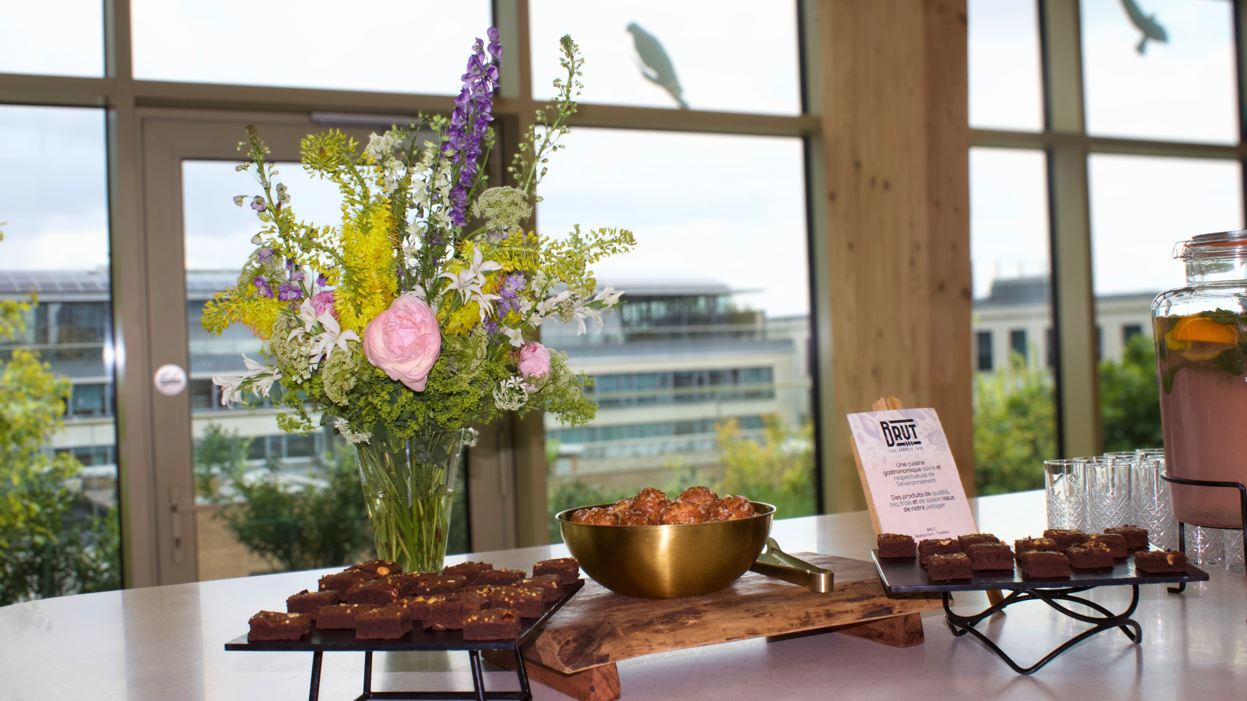 A table with a  beautiful spring bouquet of colorful flowers and a bowl of brownies, set in front of large windows in a bright room of an office building. 