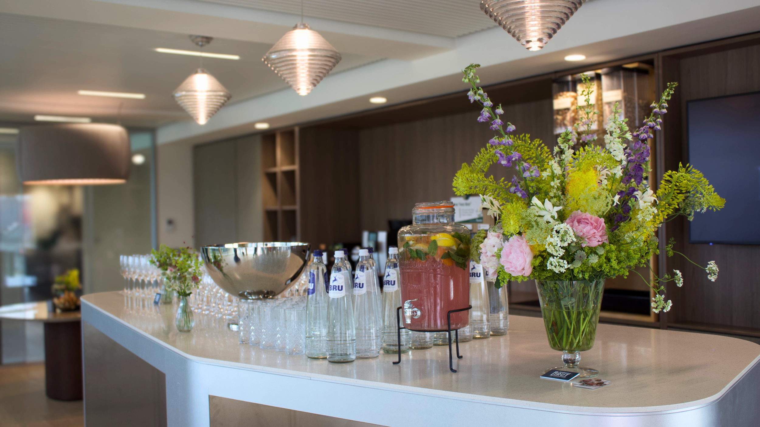 A modern reception or bar area with a white countertop, displaying a large bouquet of flowers in a glass vase, several bottles of water, and a glass jar with a pink beverage. The background features warm lighting fixtures and wooden wall panels.