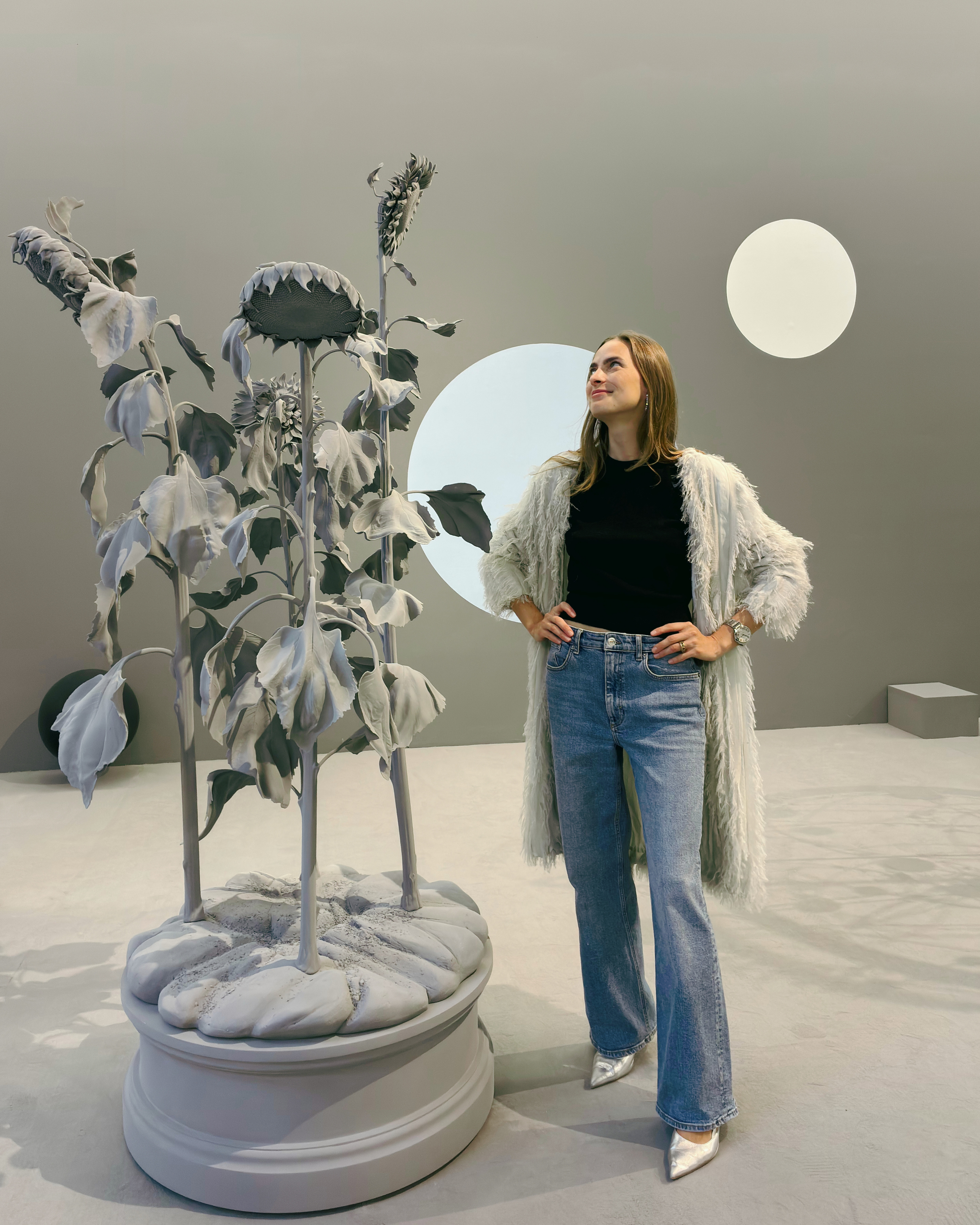 Woman with long hair wearing a grey fluffy coat, black top, and blue jeans, standing in an art installation by Hans Op De Beeck with oversized black and white sunflowers and two white circles in the background.
