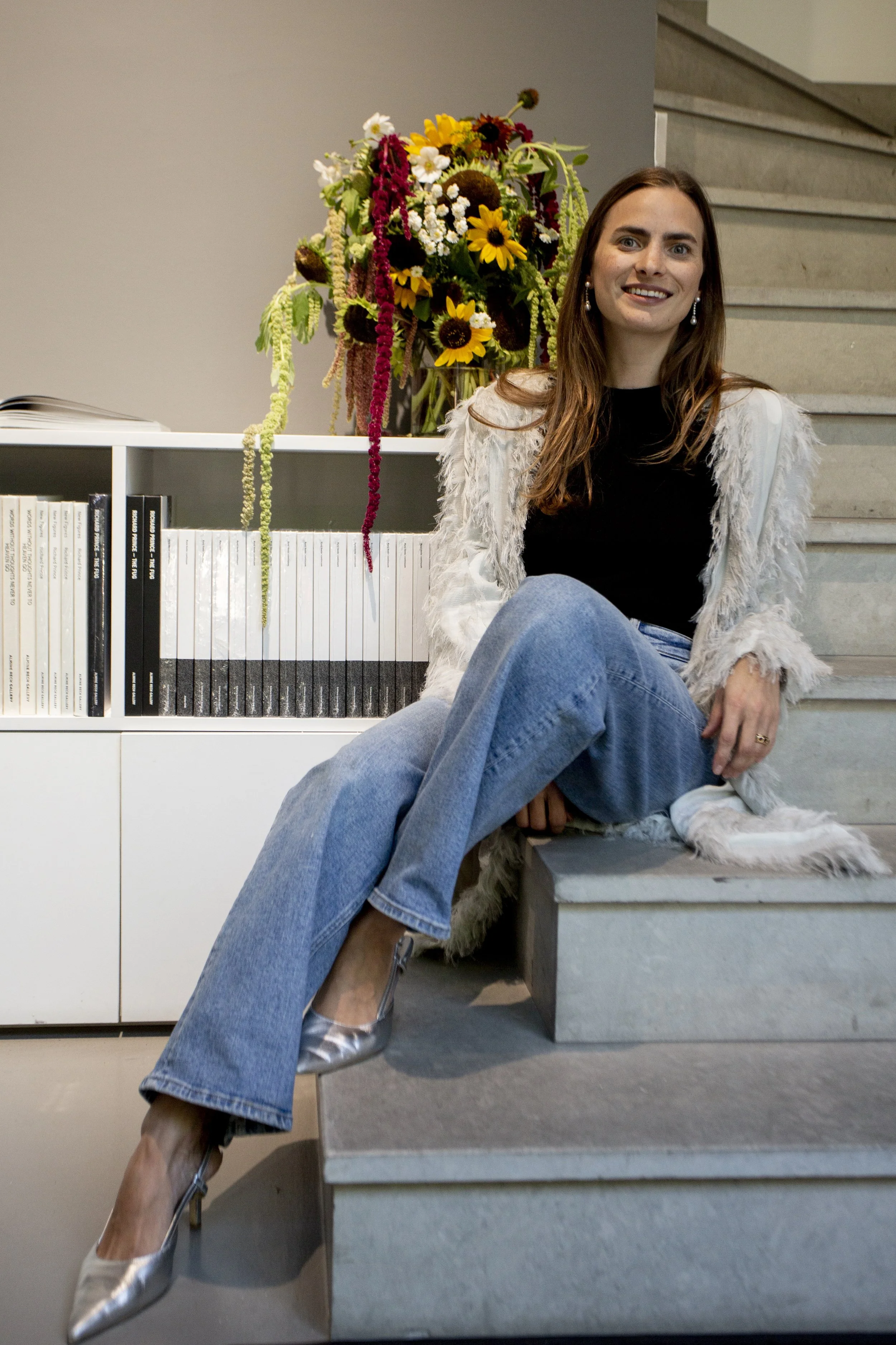 A woman sitting on a staircase, smiling, with a large bouquet of colorful flowers on a shelf behind her, wearing a white fringed jacket, black top, blue jeans, and silver heels.