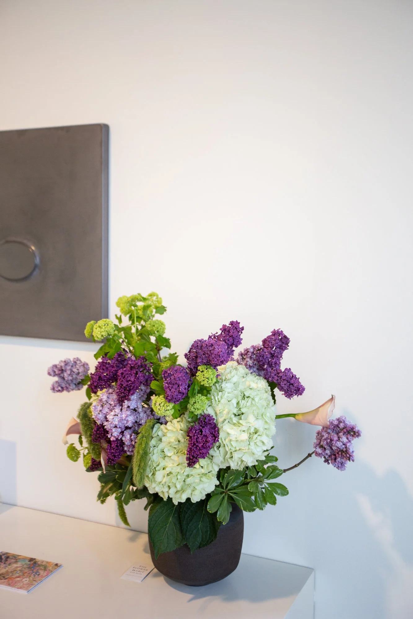 A bouquet of green hydrangeas and lilac in a dark grey ceramic vase, placed on a white table against a light-colored wall, parts of a painting are visible. 