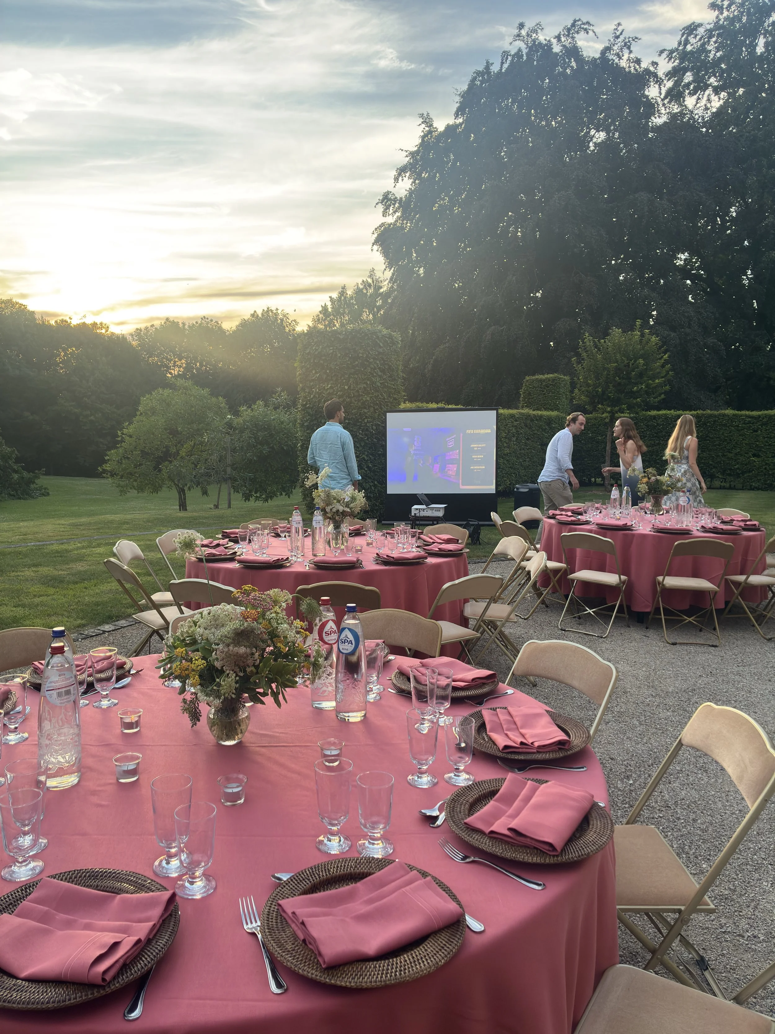 Outdoor event with round tables covered in light red tablecloths and napkins, and stunning floral centerpieces usind Daucus Carota. In the background you see large trees and a hedge, under a partly cloudy sky at sunset.