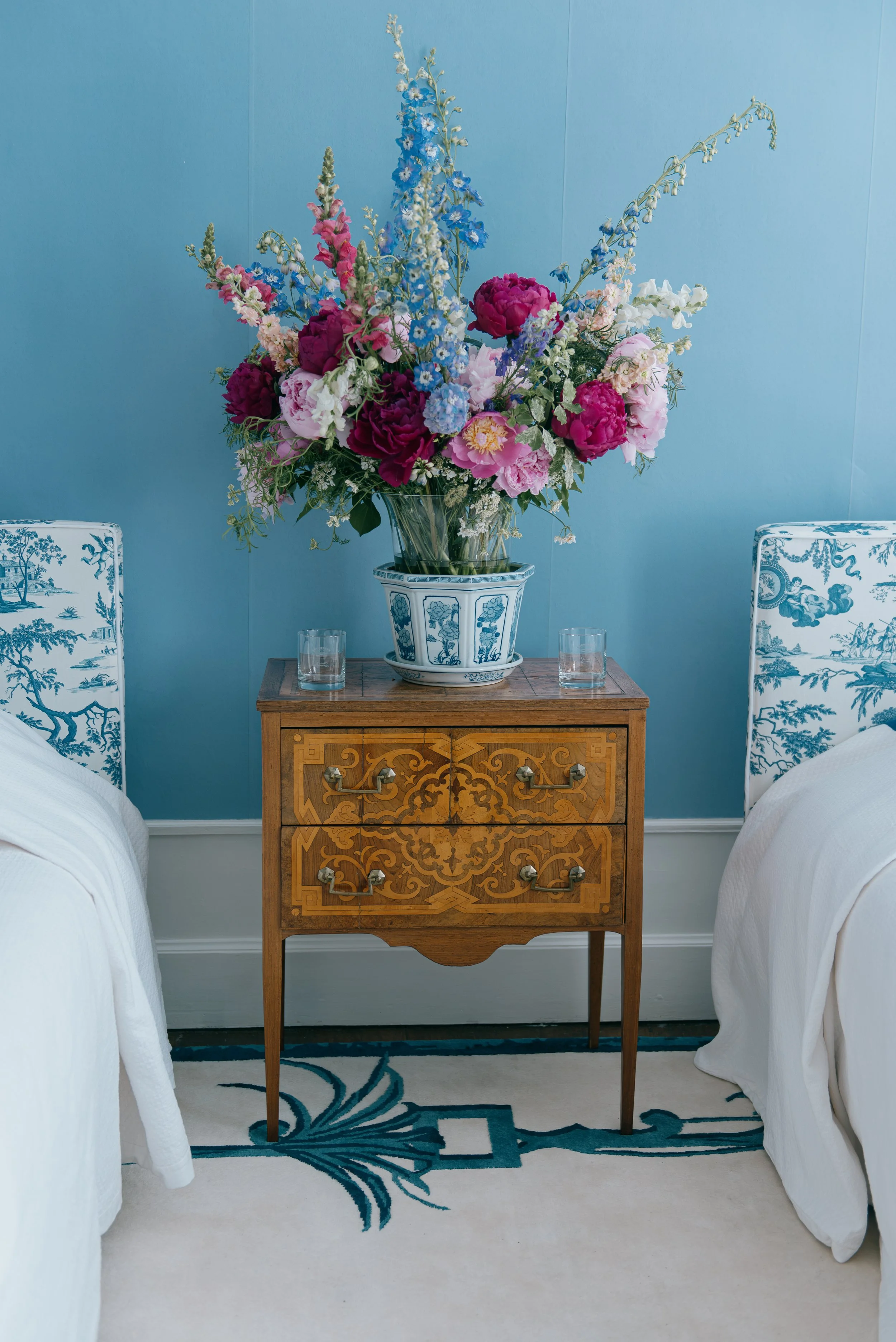 A vase with a large floral arrangement of peonies, delphinium and snapdragon, on a wooden nightstand between two elegant beds. The background wall is painted blue, and a patterned rug by Jaipur rugs is partially visible on the floor.