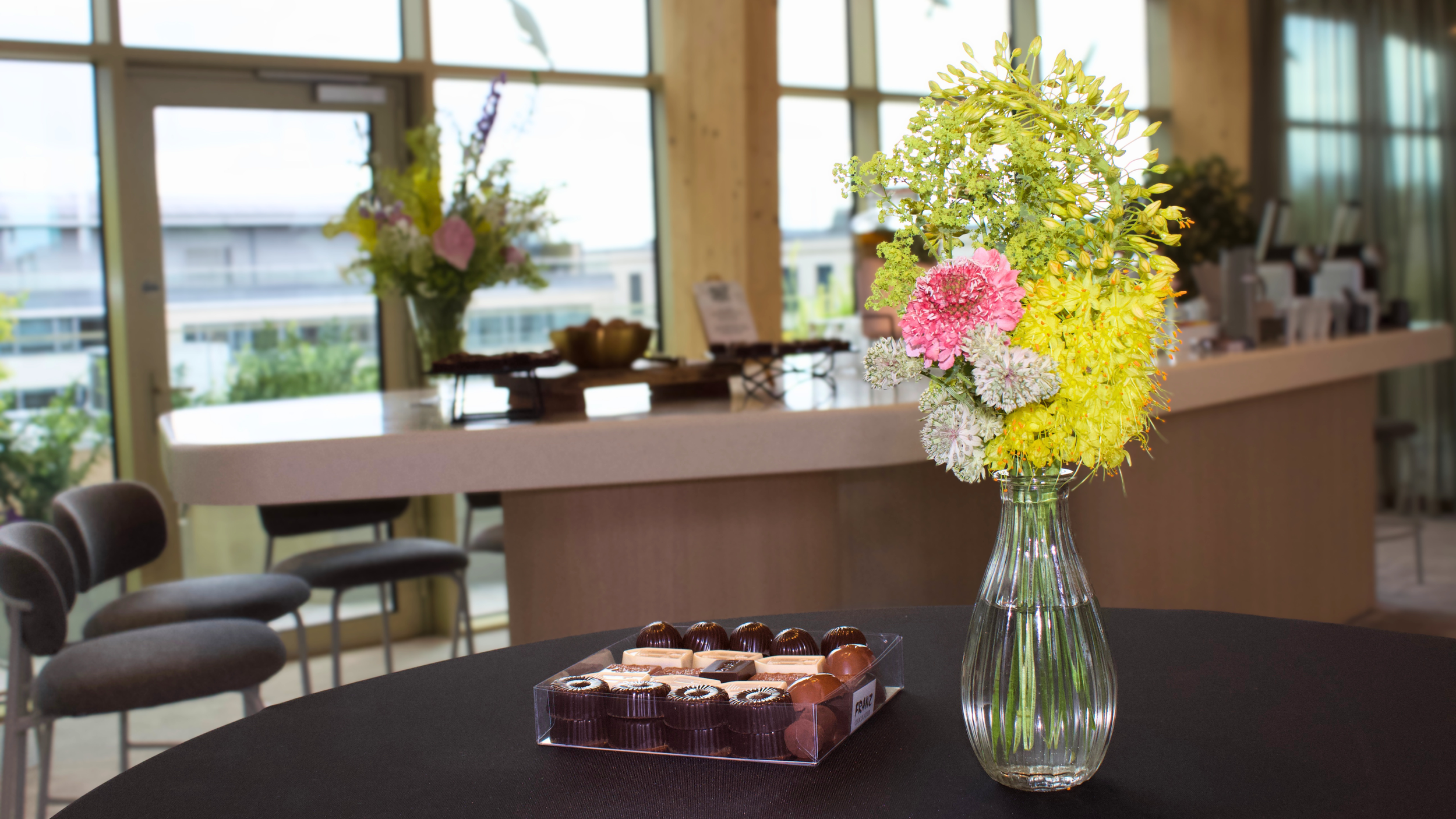 A glass vase with a colorful floral arrangement featuring pink, yellow, and white flowers on a black table, with chocolates in a clear plastic box nearby. In the background, large windows with trees outside, and a buffet or counter with more flower a