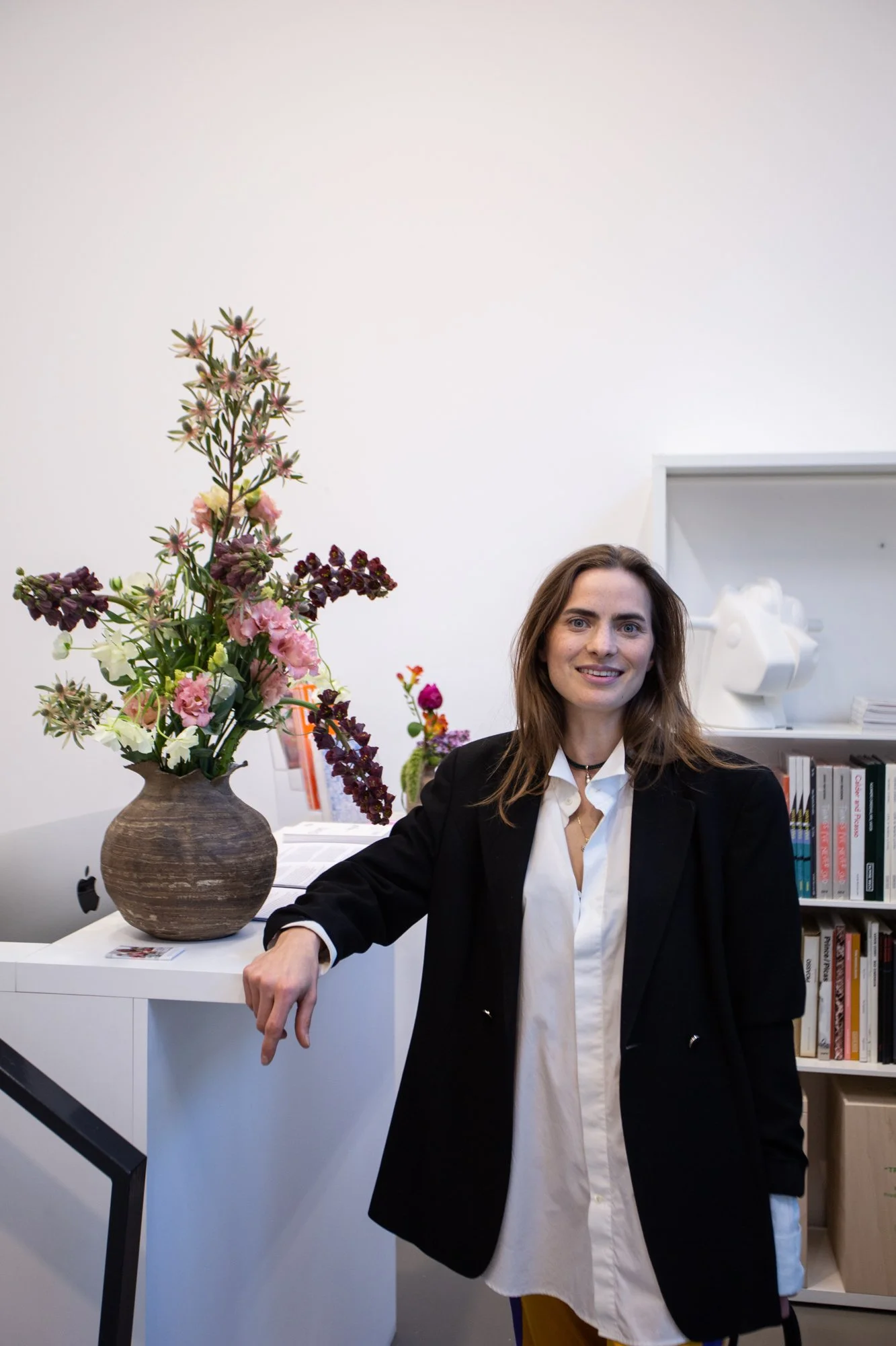 A woman with long brown hair, wearing a black blazer over a white shirt, sitting in an office surrounded by bookshelves. A large vase with an arrangement of pink, purple, white, and green flowers is on a white desk in front of her.