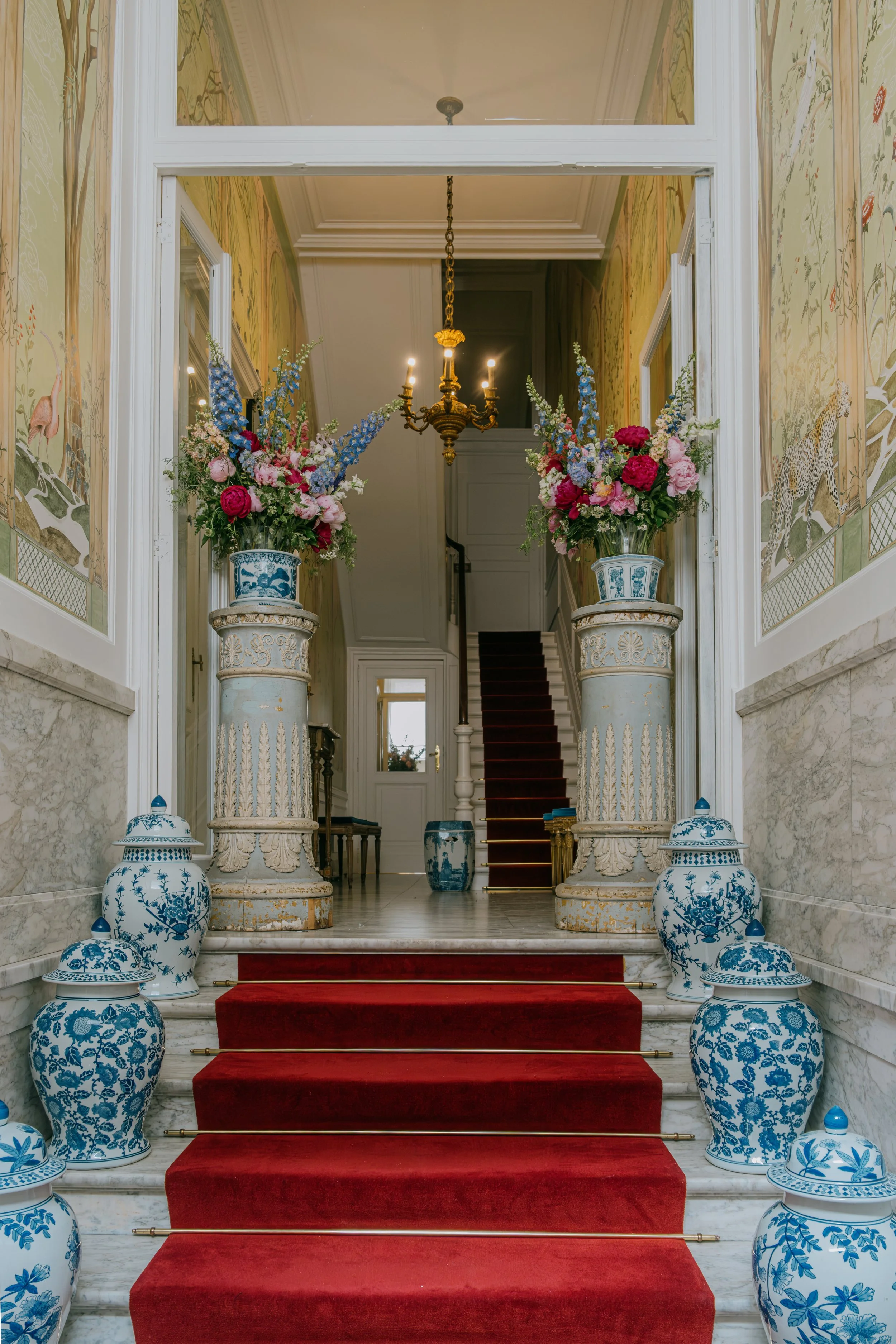 Marble staircase with red carpet runner, blue and white porcelain vases with colorful flowers, ornate wallpaper murals, a brass chandelier, and a staircase in the background.