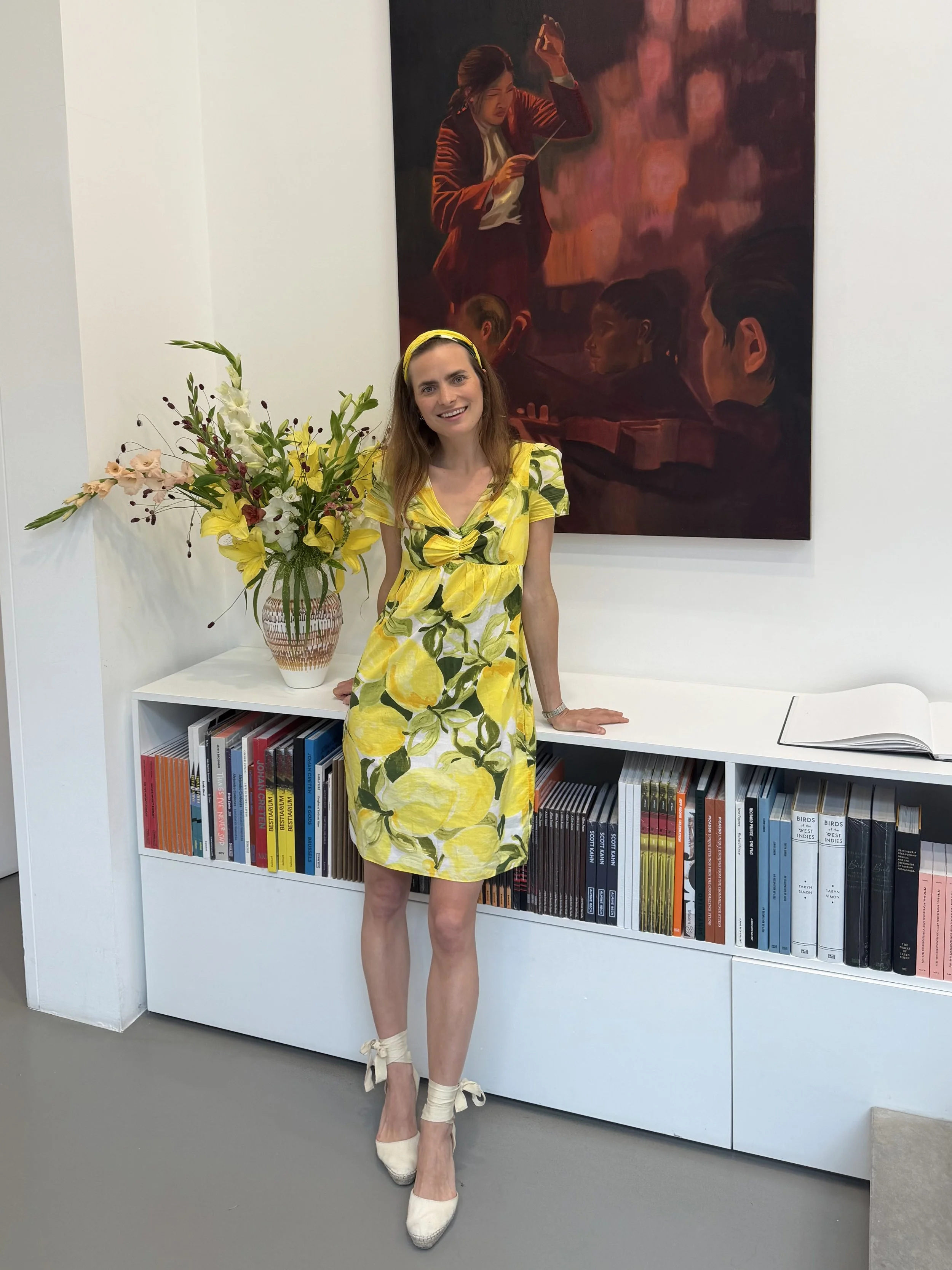 A woman in a bright yellow dress with a lemon pattern, wearing white heels and a yellow headband, standing in front of a bookshelf and a colorful painting, smiling at the camera.