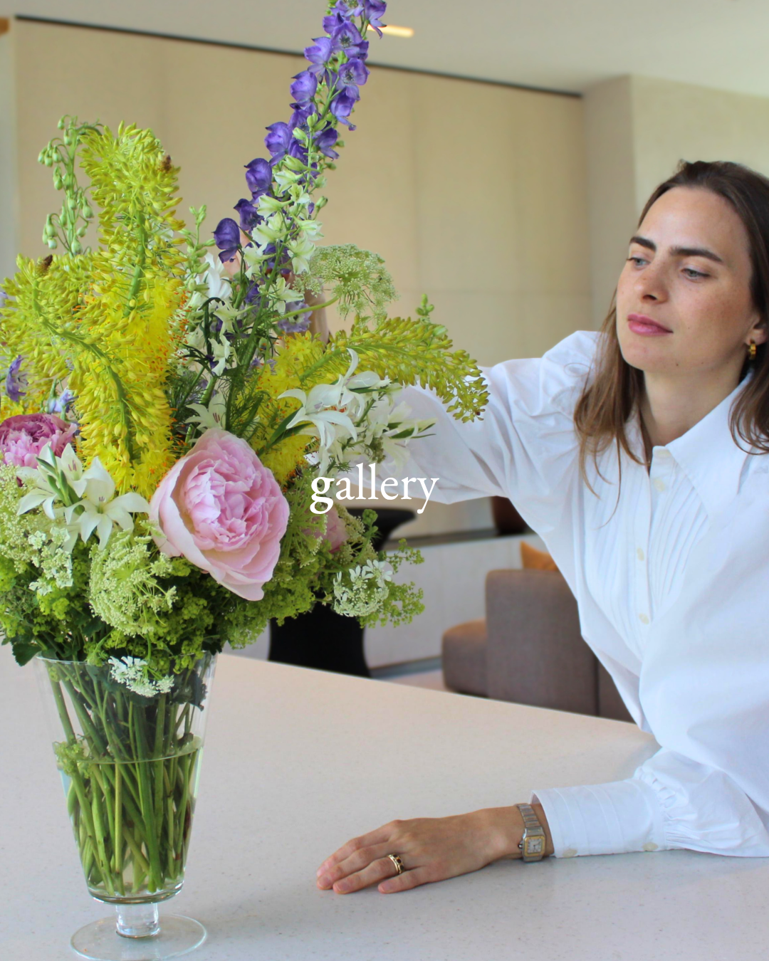 A woman arranging a large colorful floral bouquet in a glass vase, sitting at a table in an indoor setting.