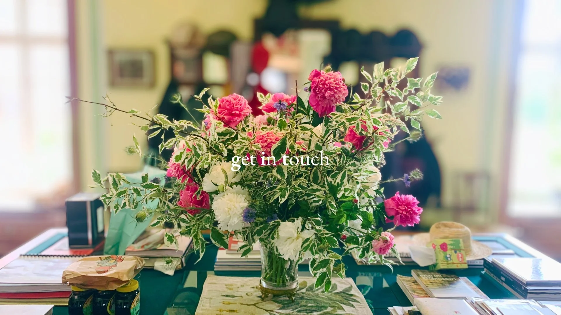 A clear glass vase filled with pink, white, and purple flowers along with green and white variegated leaves, placed on a table with books and items, with a blurred background of a room with windows and furniture, and the text 'get in touch' overlayed
