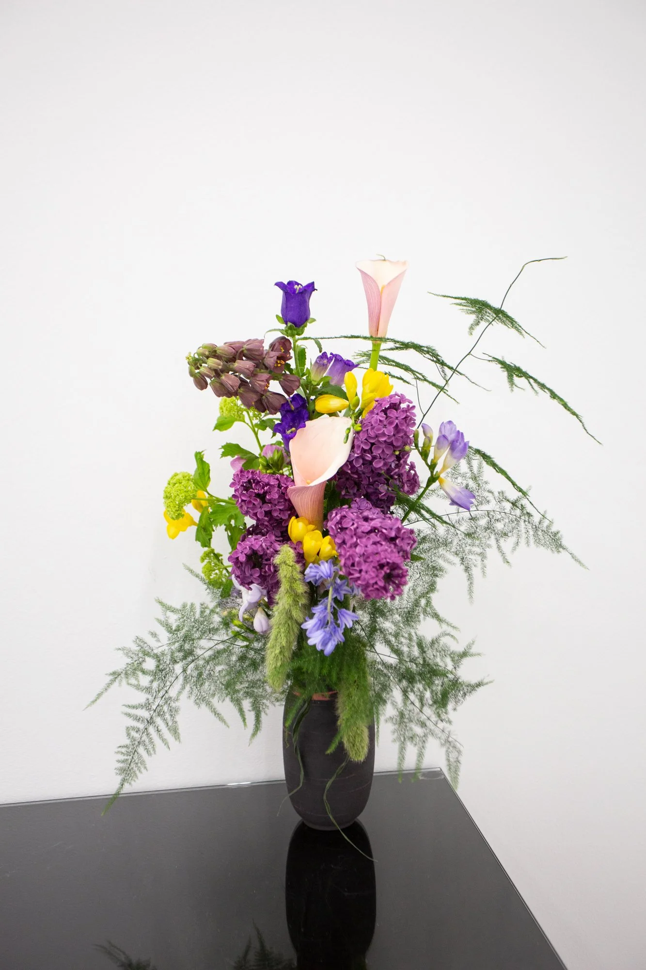A tall bouquet of assorted colorful flowers including purple, yellow, pink, and lavender blooms arranged in a black vase against a plain white wall.