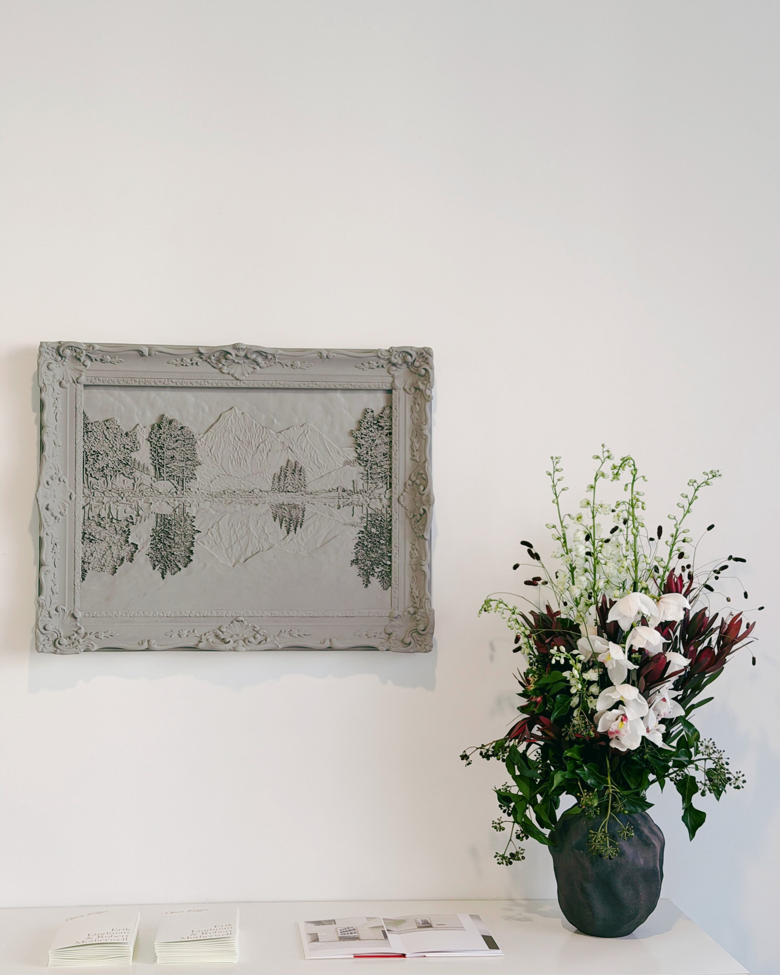 A large dark vase holding a bouquet of mostly white flowers with some dark maroon and green foliage, placed on a white surface. Behind it, mounted on a light-colored wall, is a framed black-and-white landscape carving depicting mountains, trees, and 