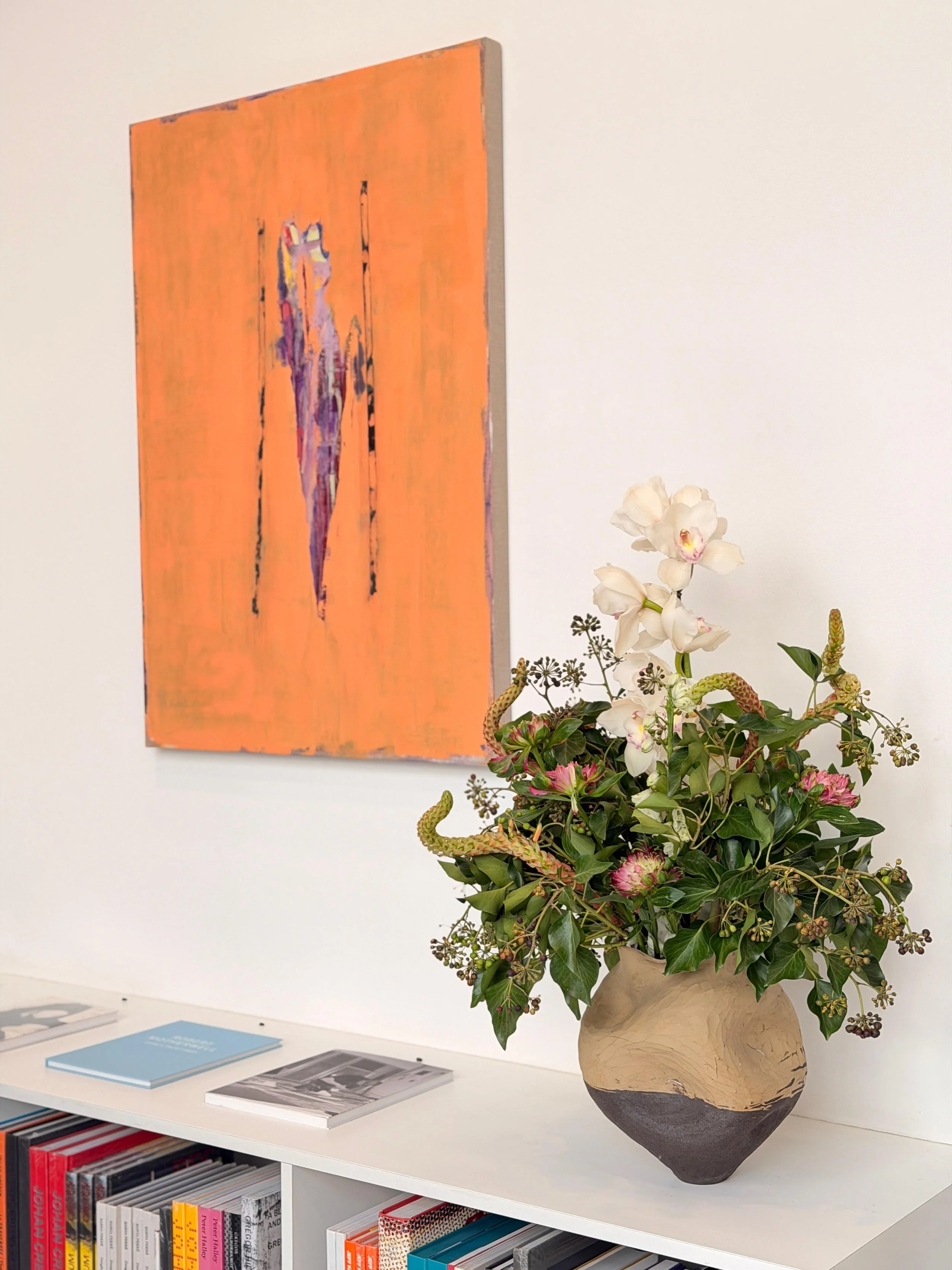A white shelf displaying books and magazines, with a large abstract painting on the wall and a beige vase with a floral arrangement including white orchids and green foliage.