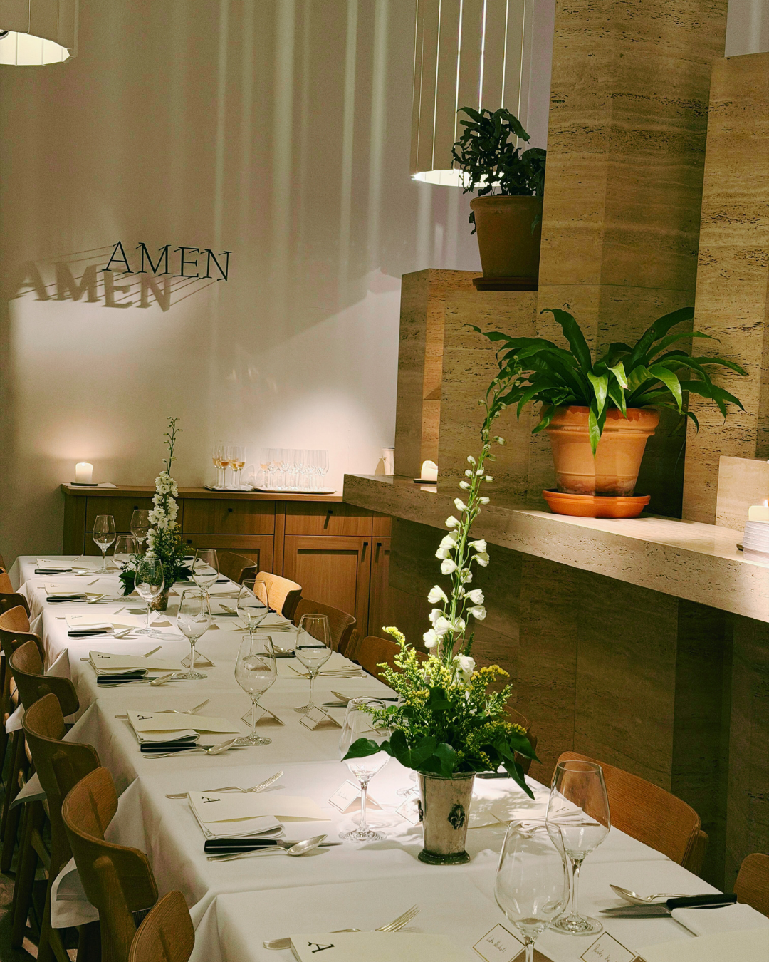 A decorated dining table set for a formal dinner with glassware, plates, napkins, and silverware, adorned with floral centerpieces, in a room with a wall-mounted plant, potted plants, and the restaurants name 'AMEN' projected on the wall