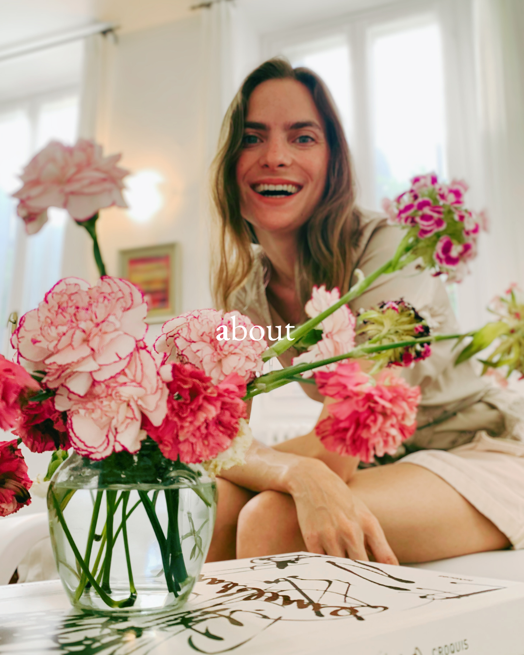 A woman with long brown hair smiling at the camera while sitting at a table with a colorful floral arrangement in a glass vase. The background shows a bright room with large windows.
