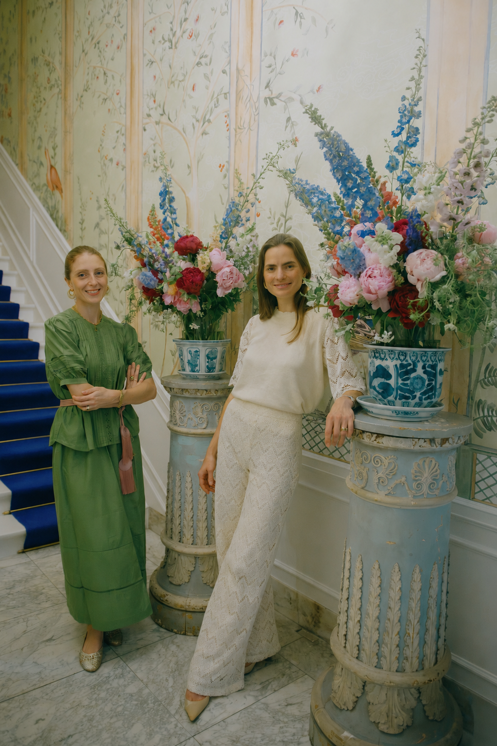 Two women standing in front of two large beautiful floral arrangements, with peonies and other spring and summer flower, in decorative blue vases, inside an elegant, painted mansion interior with marble floors and a hand painted wall paper.