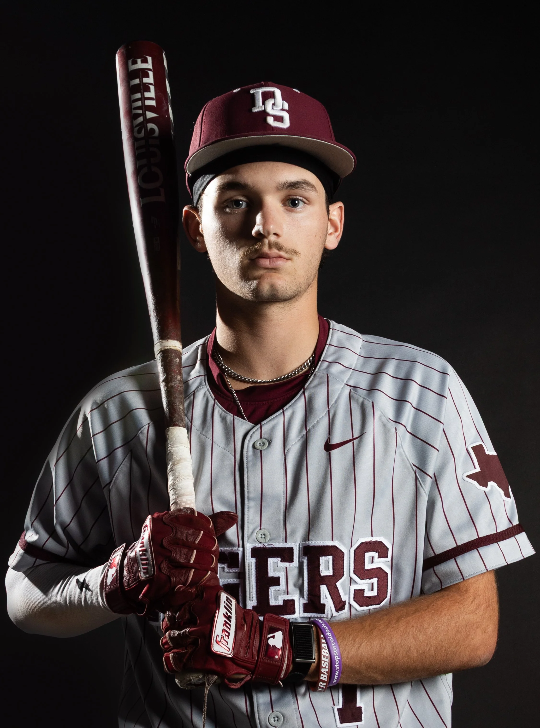 A young man in baseball uniform and cap holding a baseball bat, wearing batting gloves, against a dark background.