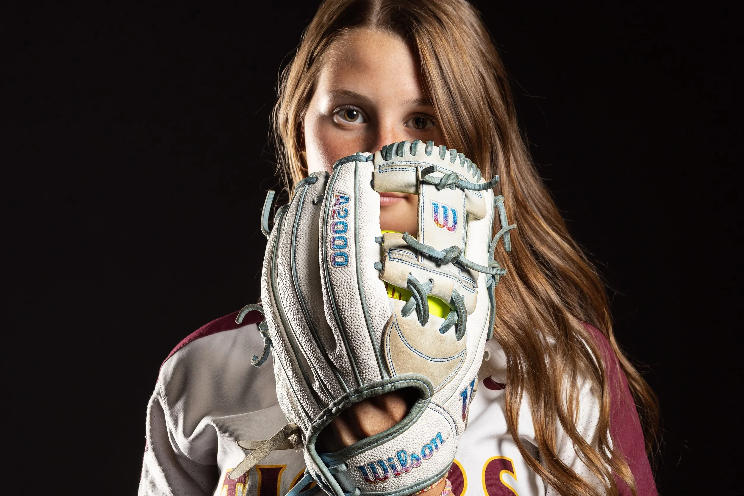 A young girl with long red hair and light skin looking directly at the camera, holding a baseball glove close to her face against a black background.