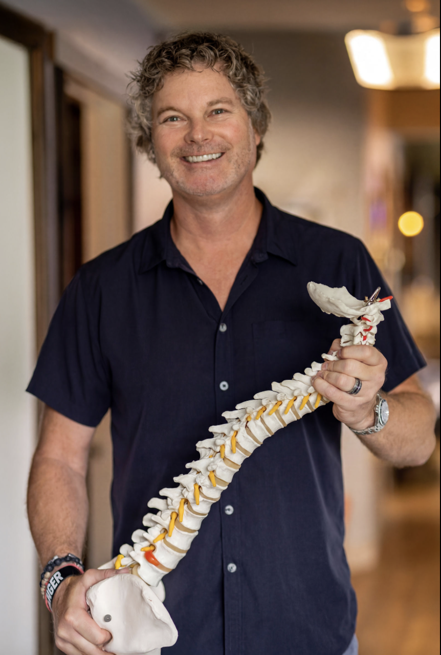A man with curly gray hair and a beard, smiling, holding a model of a human spine, standing indoors.