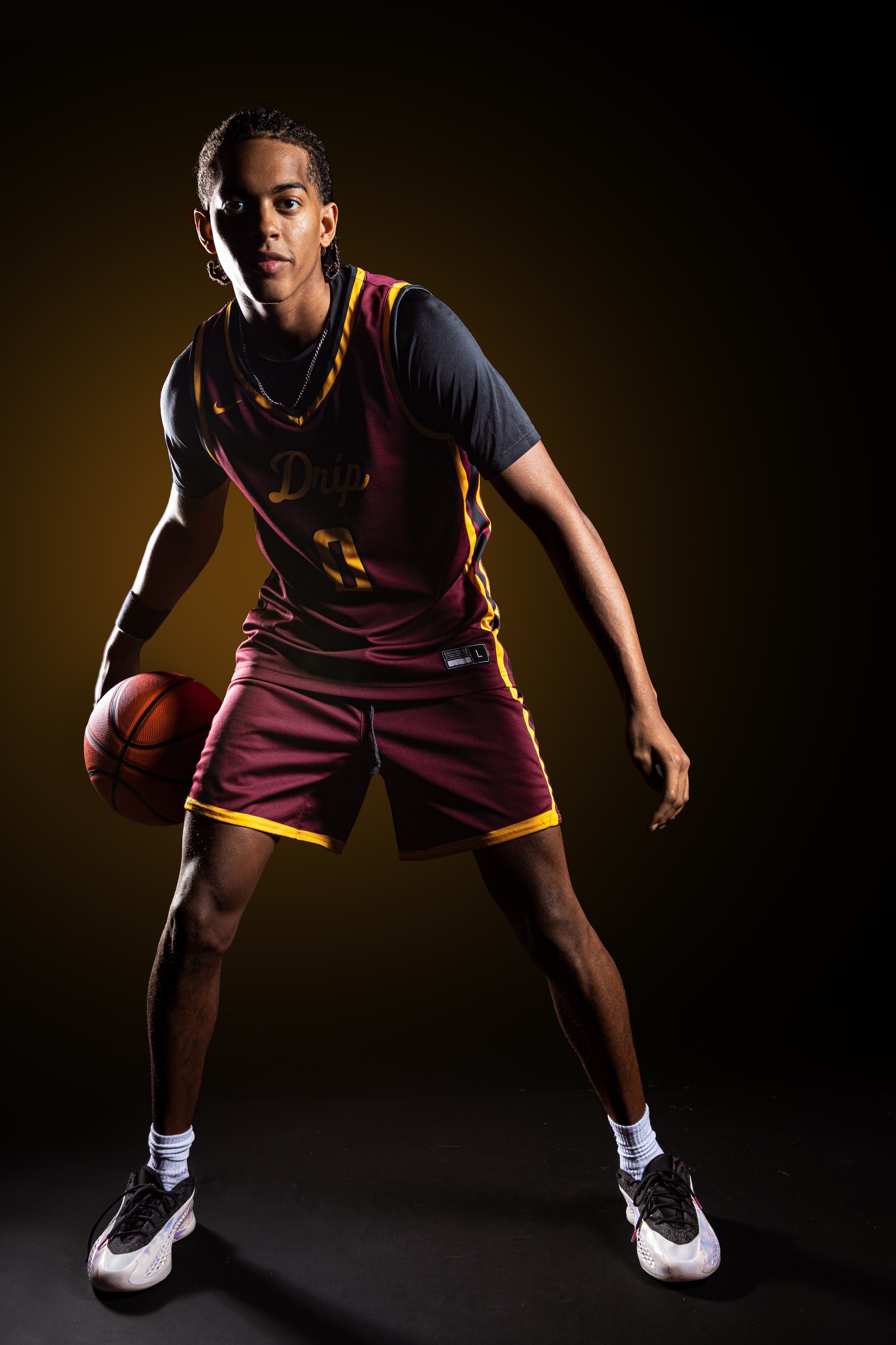 Portrait of a young male basketball player in a maroon and yellow uniform holding a basketball, standing on a black background.
