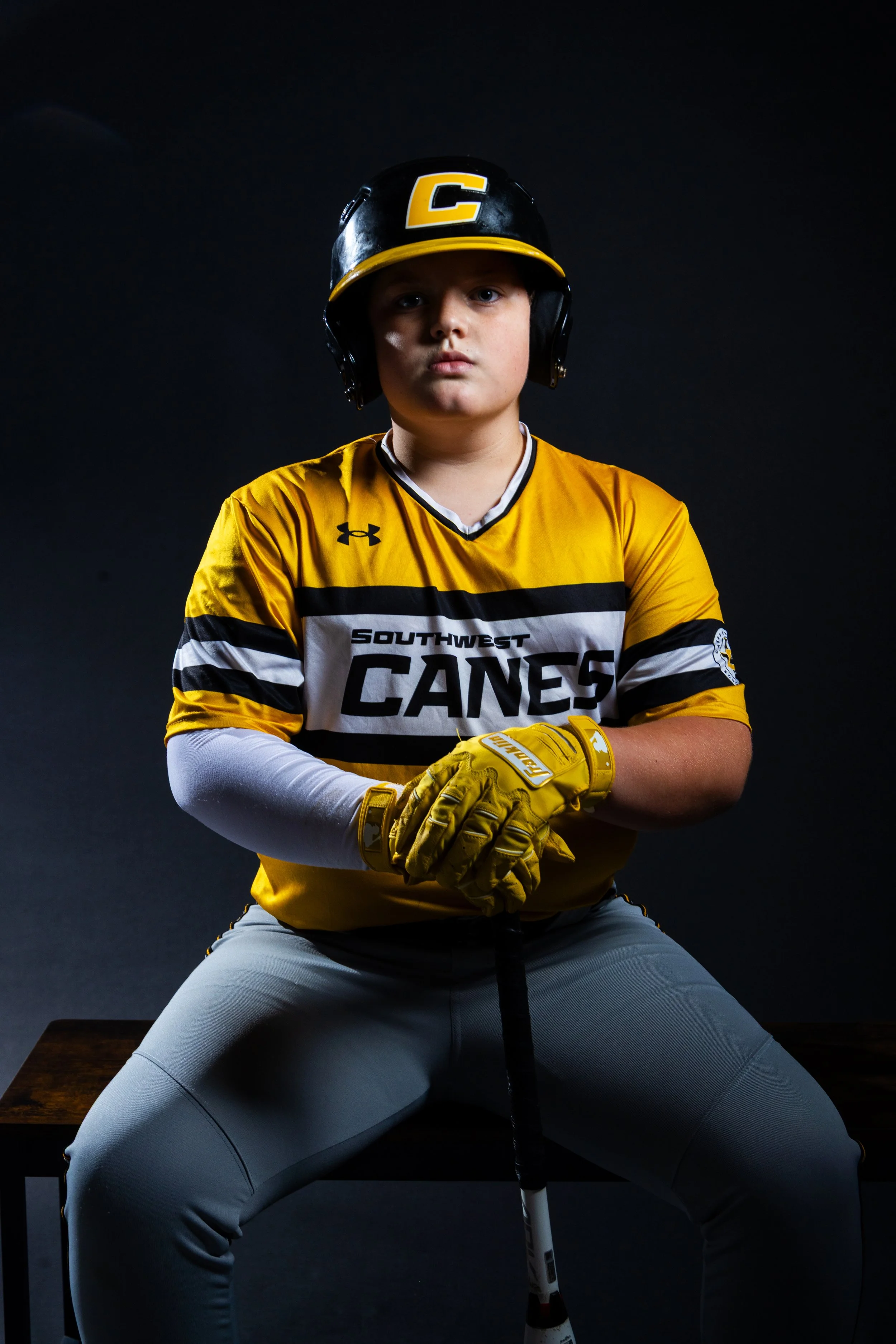 Young baseball player in yellow and gray uniform with helmet and gloves, sitting on a stool with a dark background.