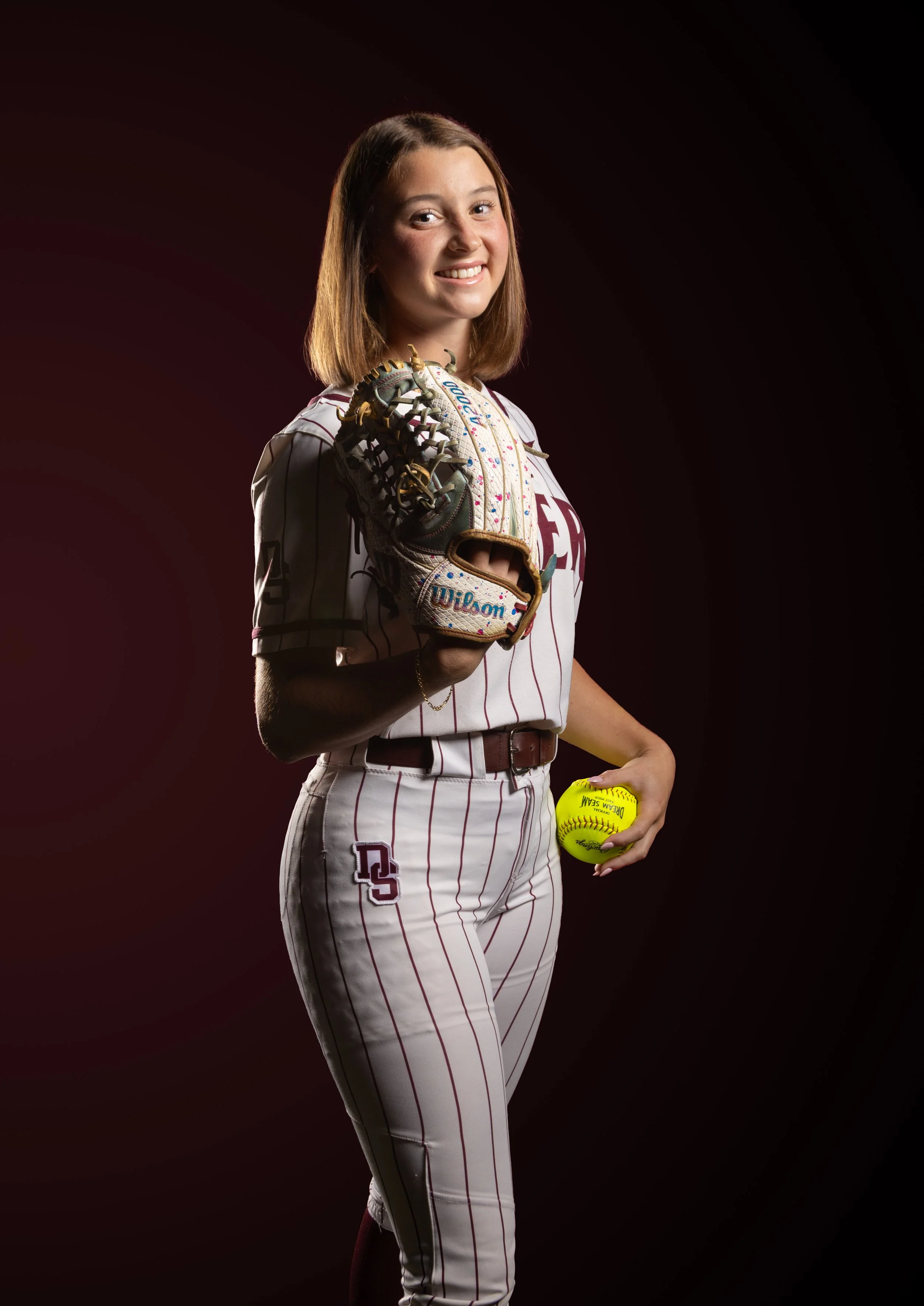 Young woman in white baseball uniform with maroon pinstripes, holding a yellow softball and wearing a baseball glove, against a dark background.