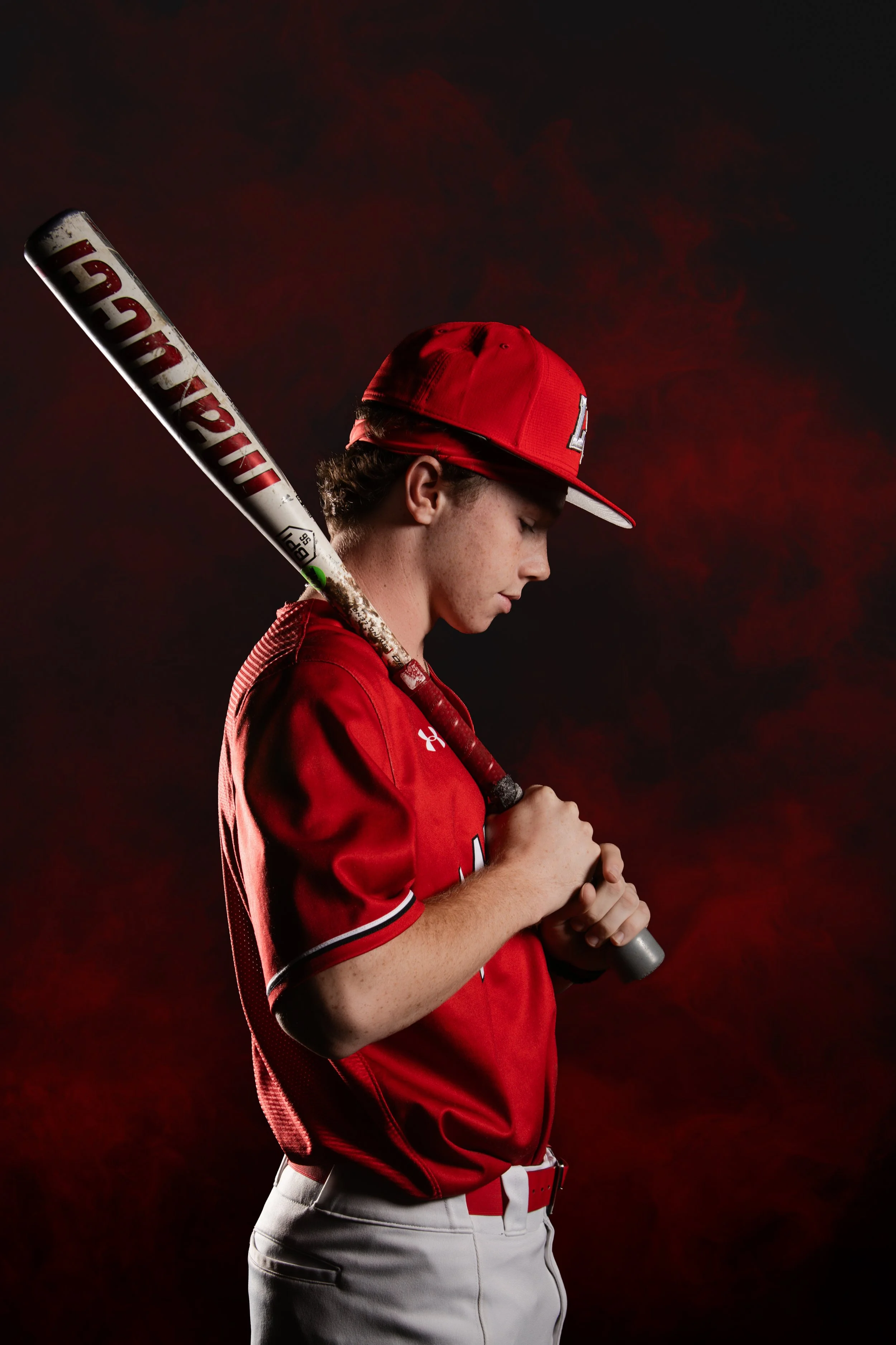A young baseball player in a red uniform and cap holding a bat against a dark, smoky background.