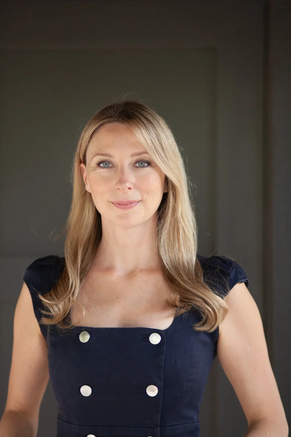 A woman with blonde hair and blue eyes smiling softly, wearing a navy blue dress with white buttons, standing against a gray background.