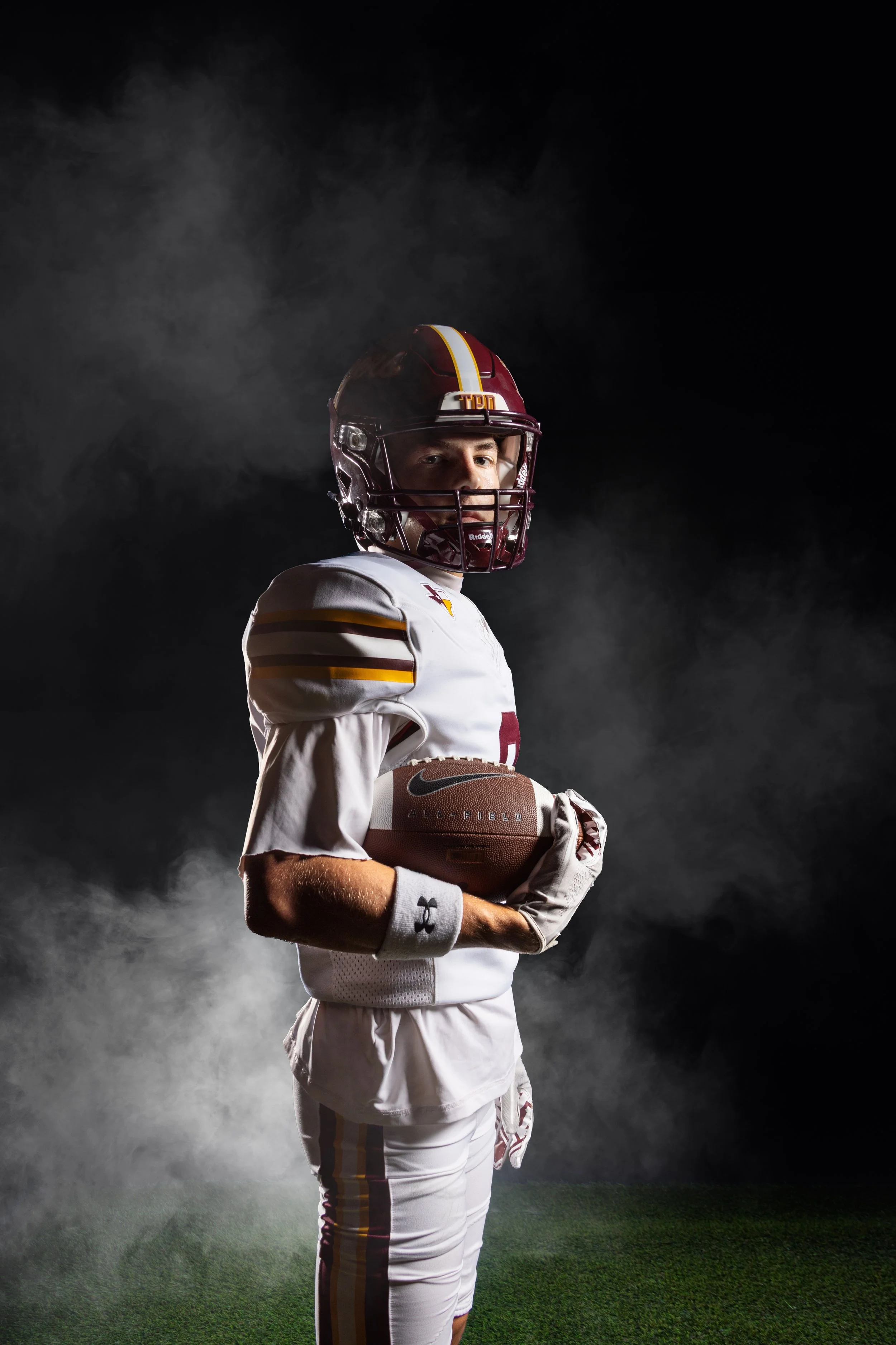 A football player in a white uniform with maroon and gold accents stands on a football field, holding a football, with a dark background and fog surrounding him.