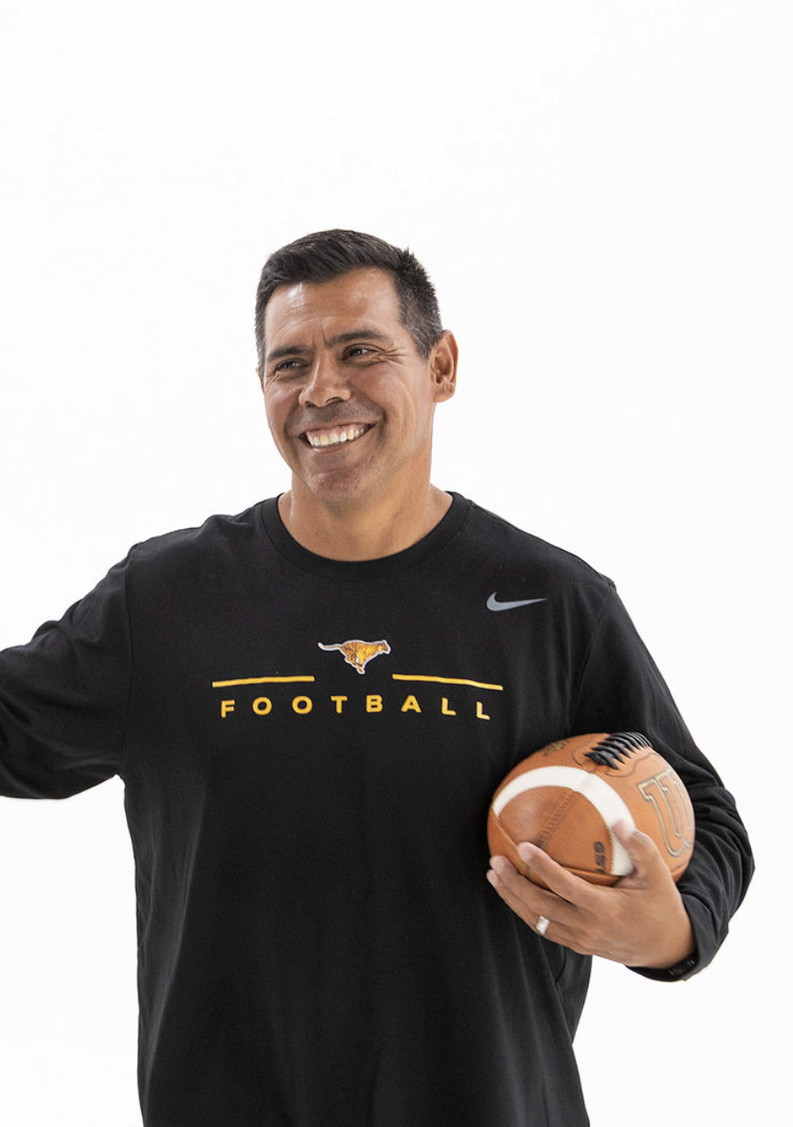 Man smiling and holding a football, wearing a black sports shirt with "FOOTBALL" and a logo of a longhorn. White background.