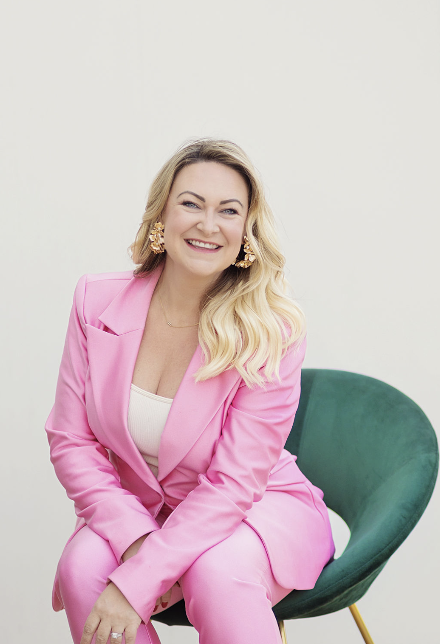 A woman with long blonde hair wearing a pink blazer and pink pants, sitting on a green chair, smiling at the camera against a plain white background.