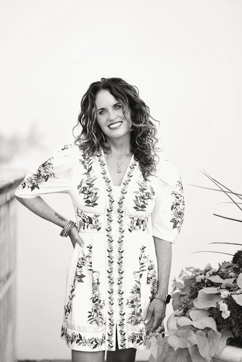 Black and white photo of a smiling woman with curly hair wearing a floral embroidered dress, standing outdoors near a railing and potted plants.