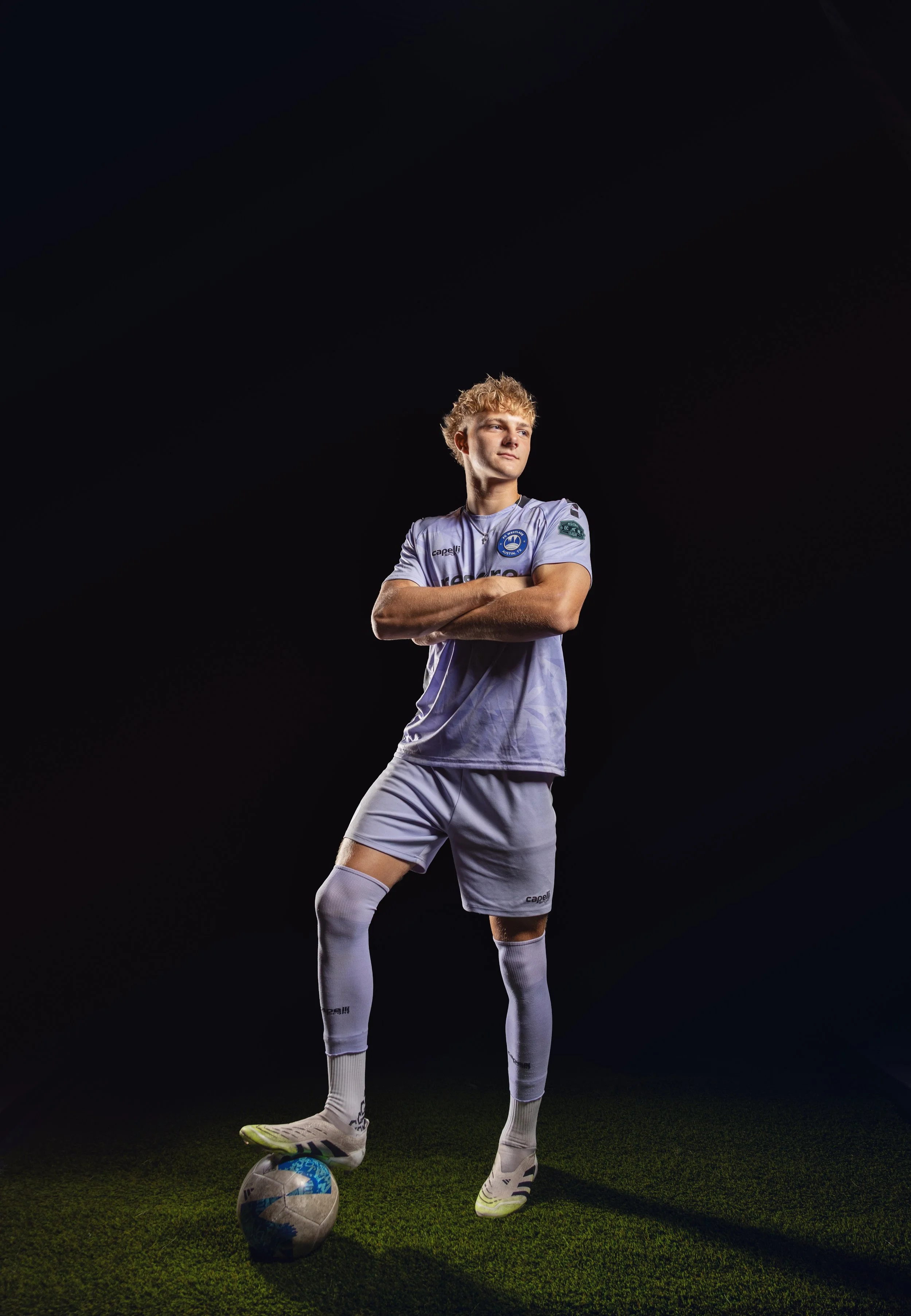 A young male soccer player in a white uniform stands with crossed arms, one foot on a soccer ball, under dark lighting with a black background.
