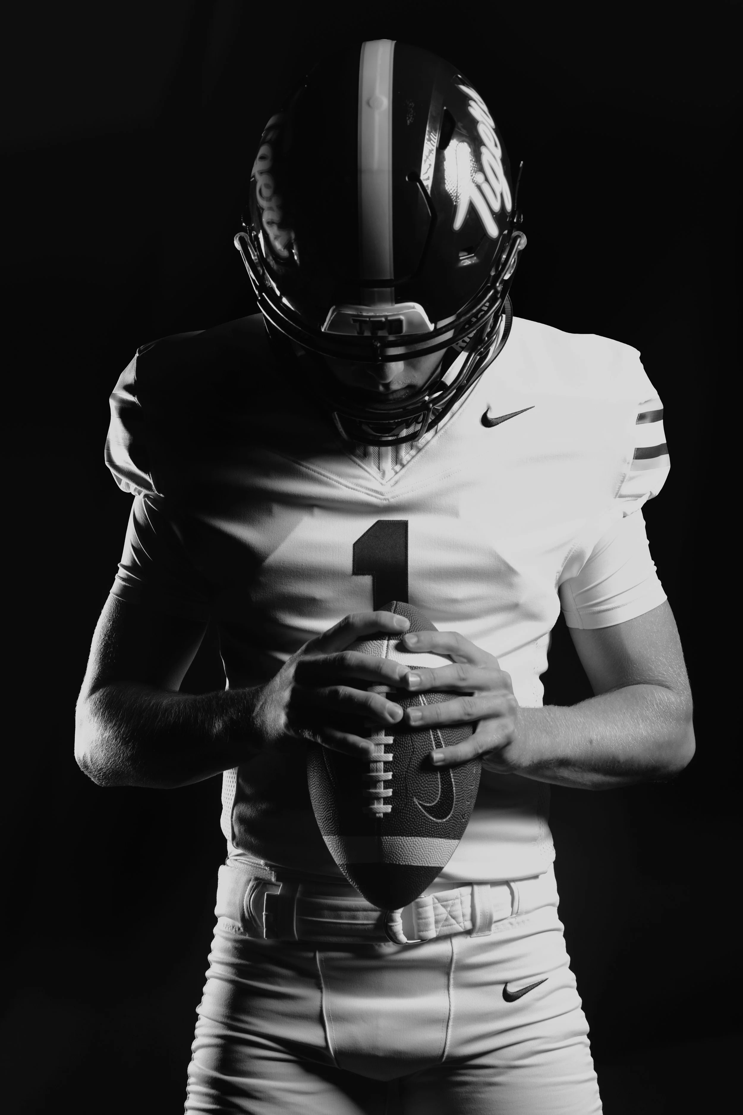 Black and white photo of a football player wearing a helmet and uniform, holding a football with both hands, against a dark background.