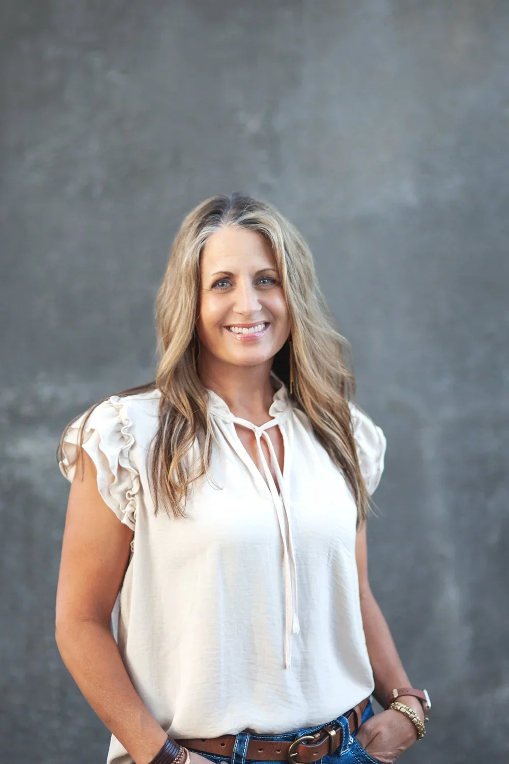 A woman with light brown, wavy hair, smiling, wearing a cream-colored blouse with ruffled sleeves, jeans, a brown belt, and bracelets, standing against a grey background.