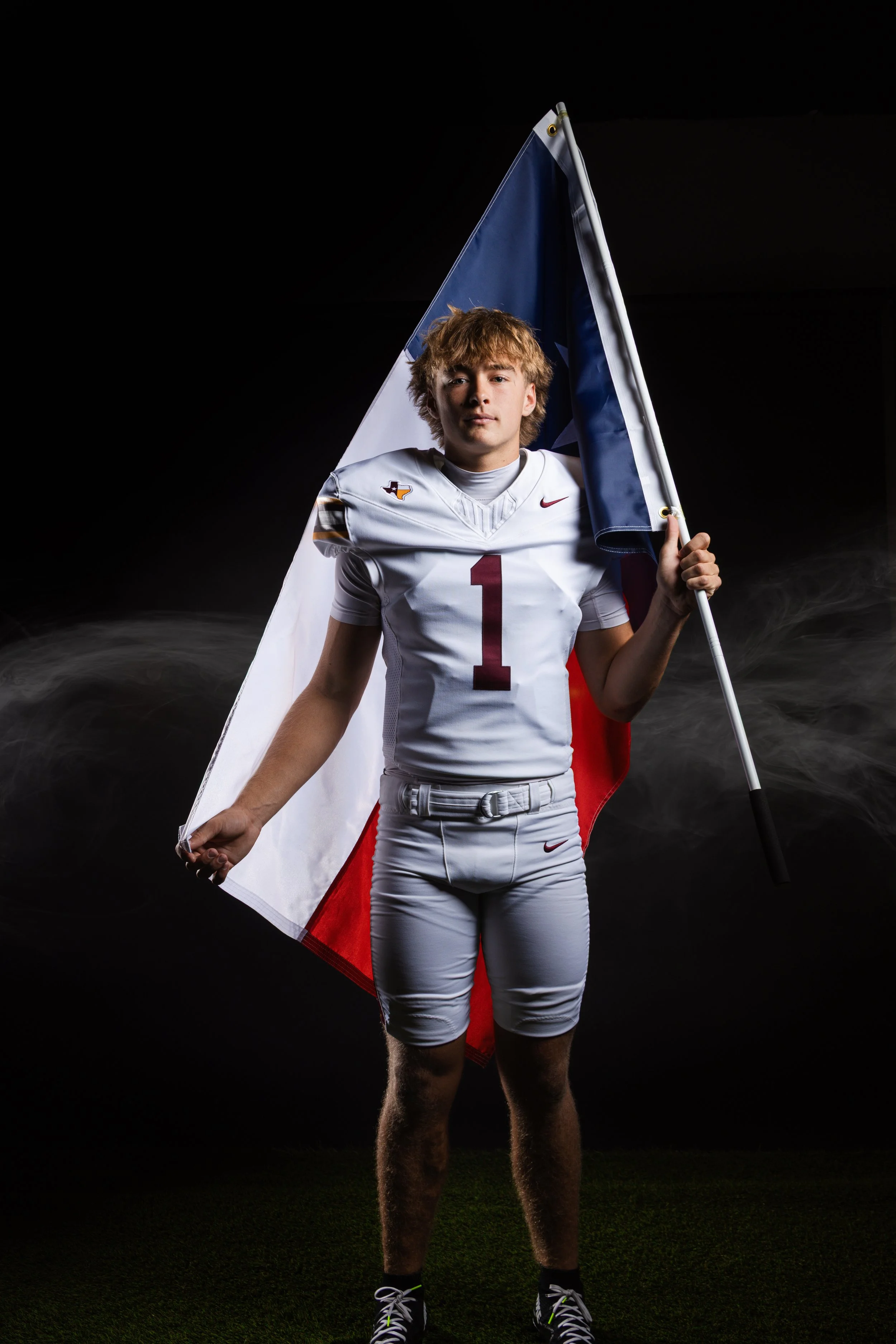 Young male football player in athletic uniform holding a American flag, with a black background and a spotlight emphasizing his determined expression.
