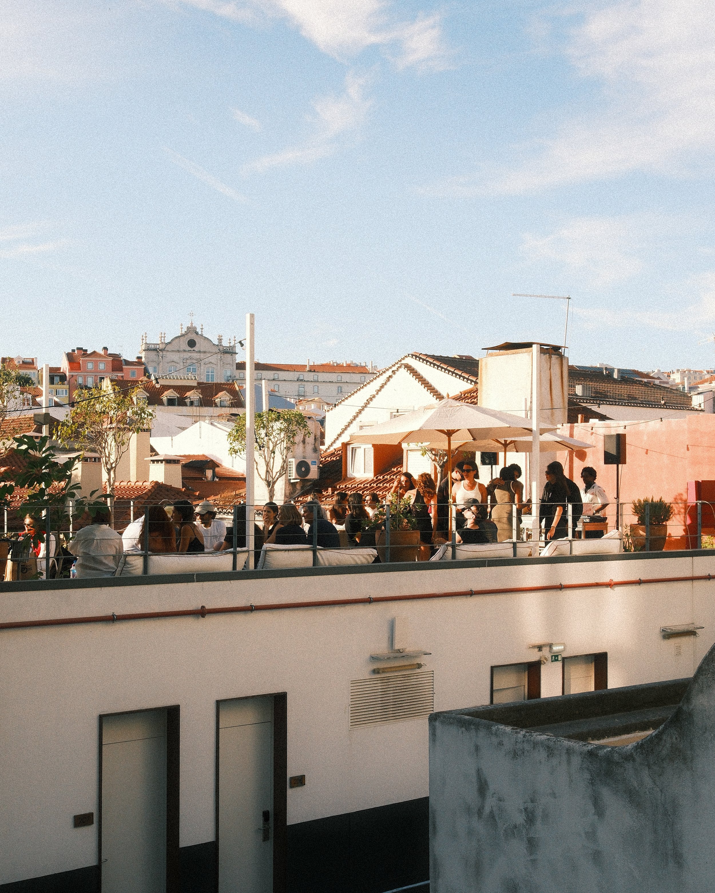 People gathered on a rooftop terrace with umbrellas, overlooking Lisbon's cityscape with rooftops and historic buildings under a blue sky with wispy clouds.