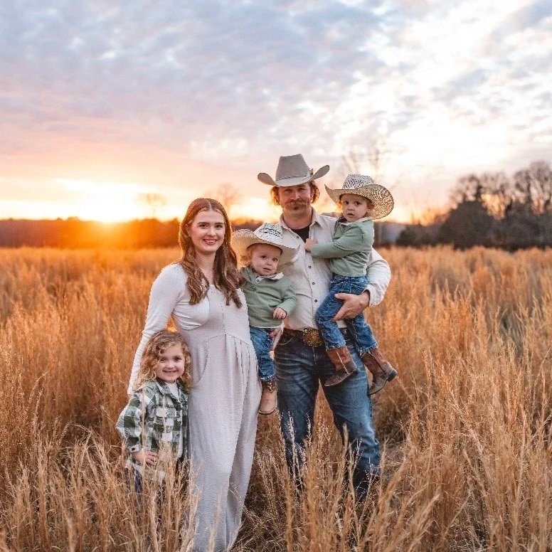 A family of five standing in a grassy field during sunset, with the parents holding two young children, all dressed in country-style attire and cowboy hats.