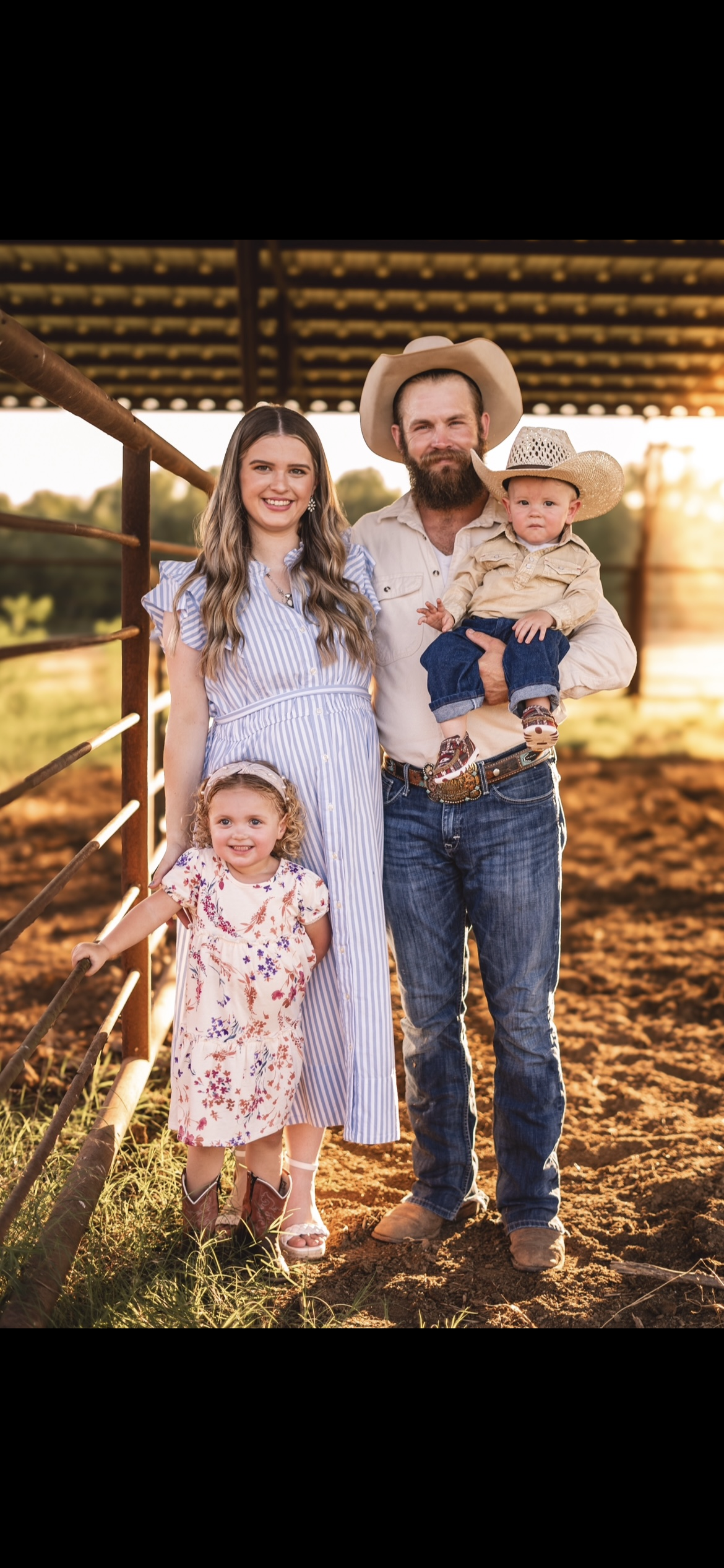 A family of five posing outdoors on a farm during sunset, including a woman in a blue striped dress, a man with a beard and cowboy hat holding a young boy, a young girl in a floral dress, all standing on dirt by a metal fence with a barn in the background.