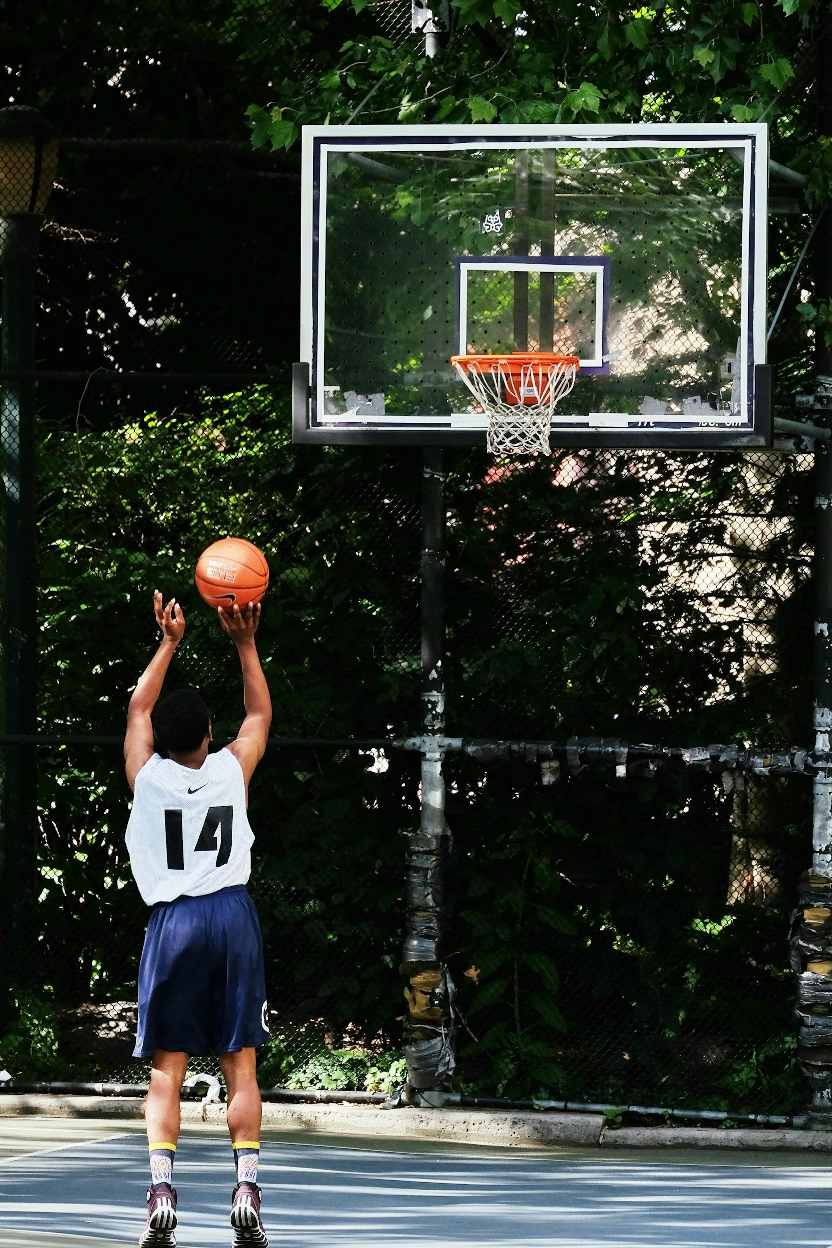 A person wearing a white jersey with the number 14 shooting a basketball on an outdoor court with a black chain-link fence and green trees in the background.