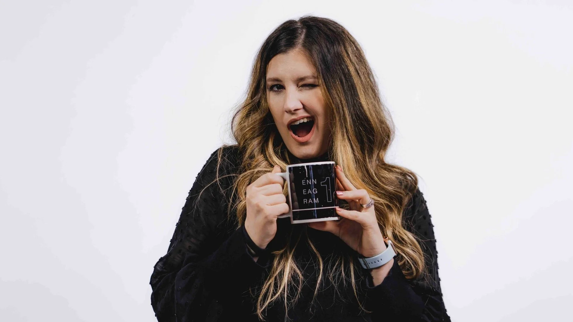 Alex Fittin is an Enneagram Coach for mission-focused organizations. She is holding a black and white coffee mug with the words 'ENNEAGRAM 1' printed on it, and she is making a surprised or excited expression with one eye closed and her mouth open.