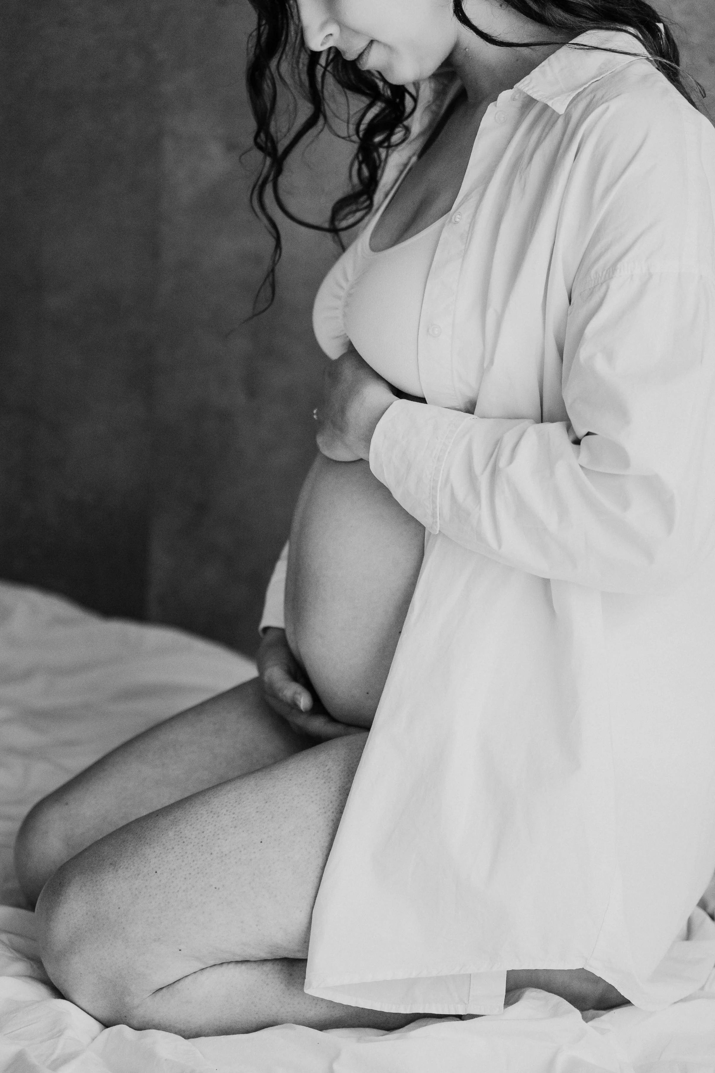 Une femme enceinte en position assise, portant un chemisier blanc, regardant tendrement son ventre, en noir et blanc.