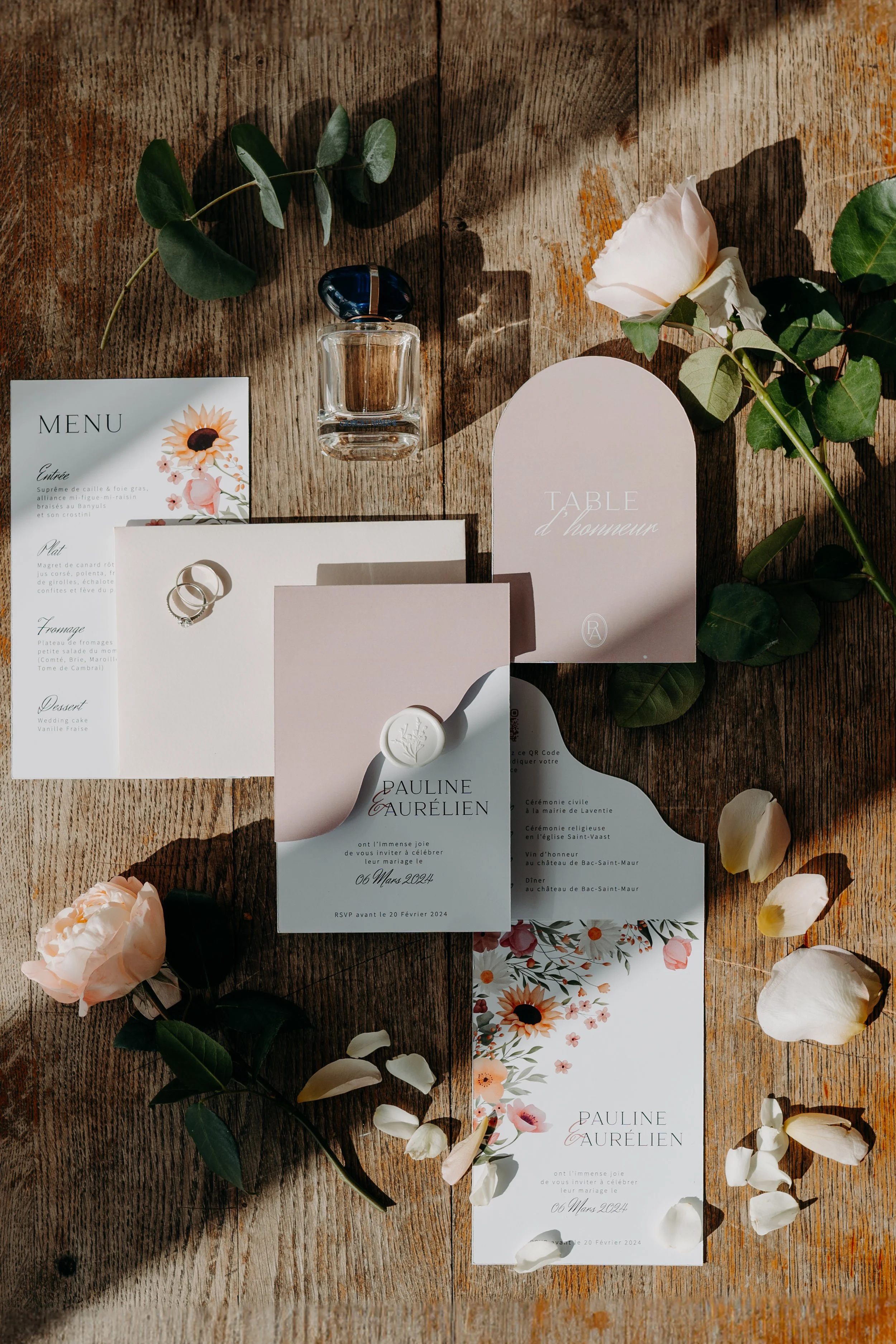 Invitation de mariage avec fleurs, feuilles, un verre de parfum, des Roses, des pétales, des cartes de remerciement et des alliances sur une table en bois. Matetmoica
