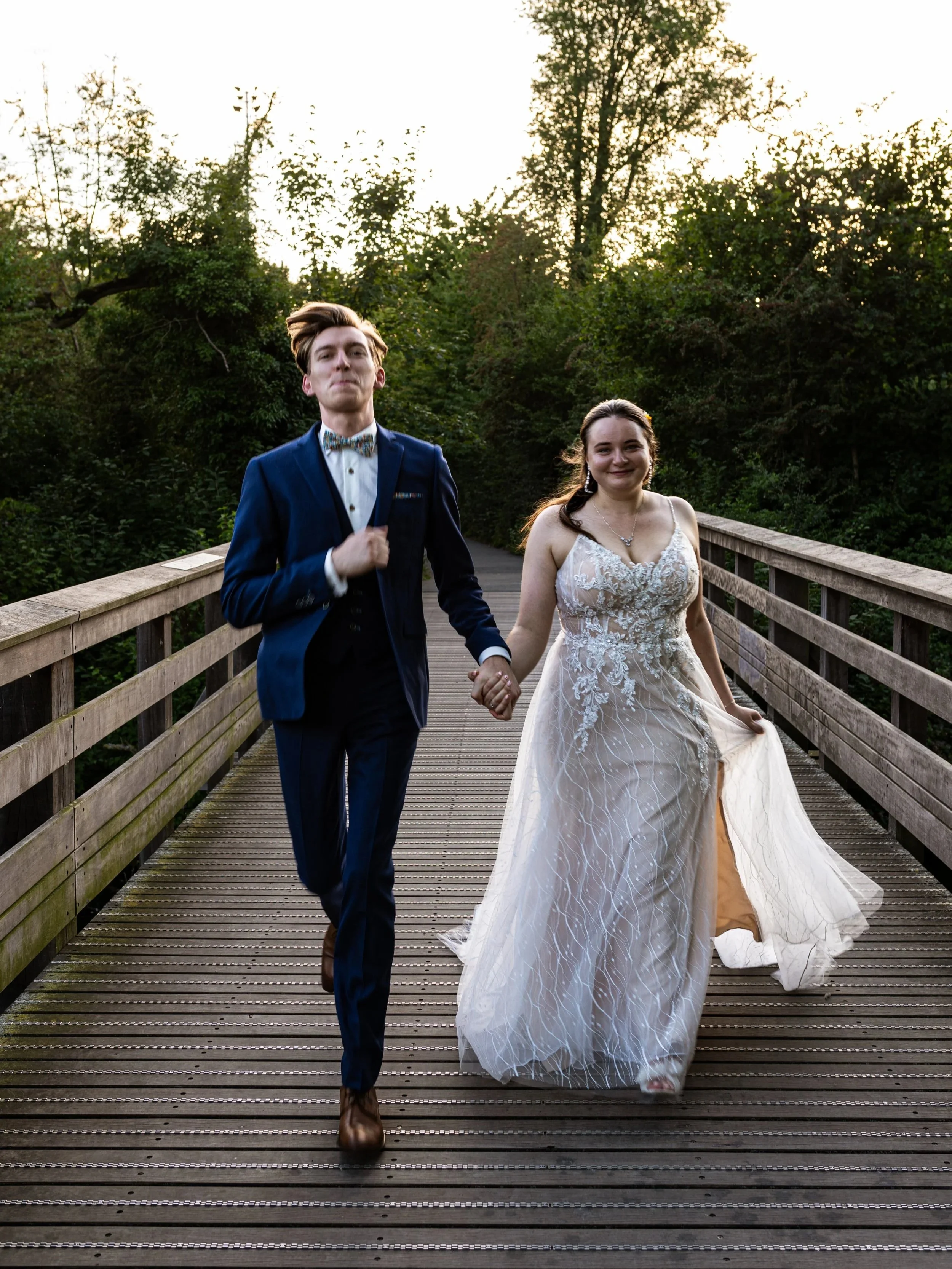 Un couple en tenue de mariage marche sur un pont en bois entouré de verdure, la lumière du soir illumine la scène.