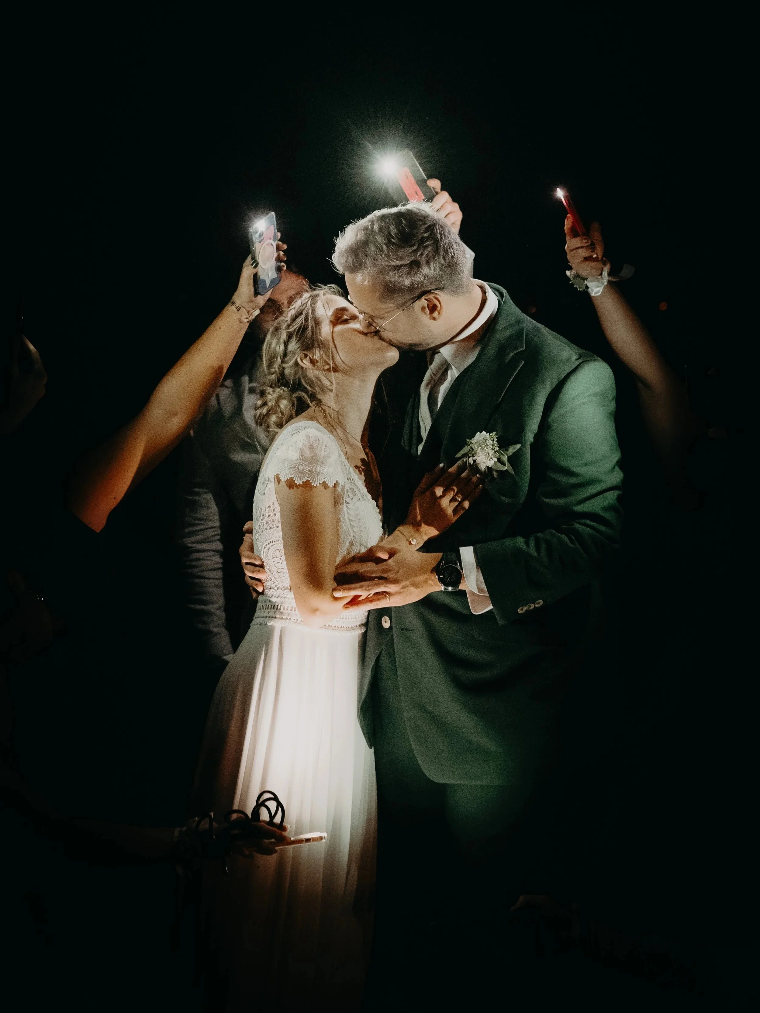 Un couple emploie une bouchée de baiser lors de leur mariage, entouré d'invités tenant des téléphones pour prendre des photos, dans un cadre nocturne.