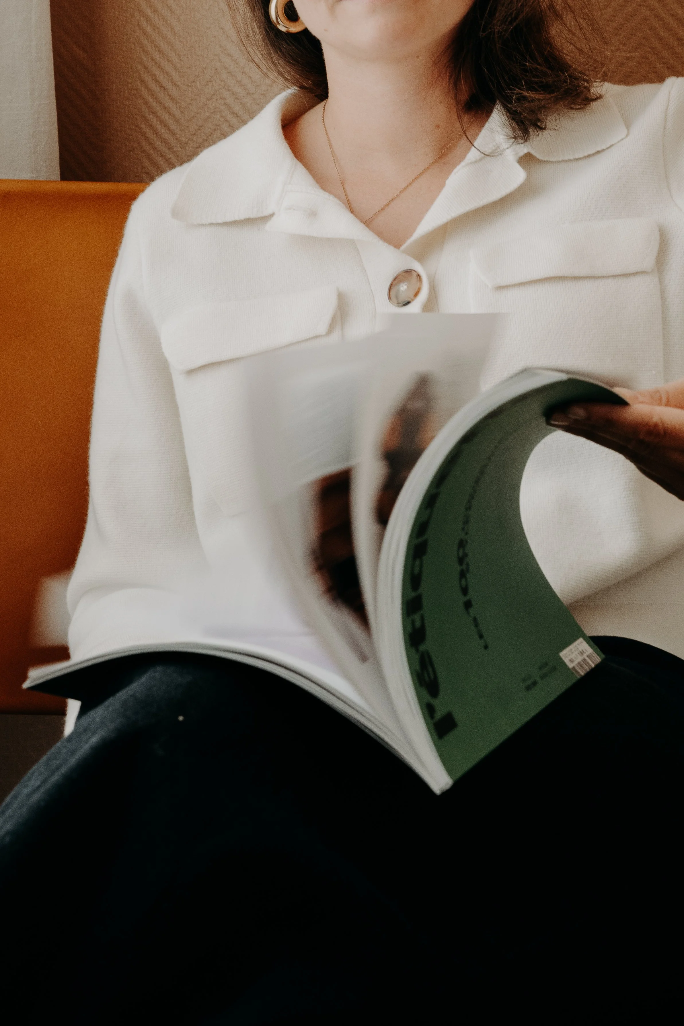 Une femme lit un magazine, portant une chemise blanche avec des boutons et une chaîne en or.