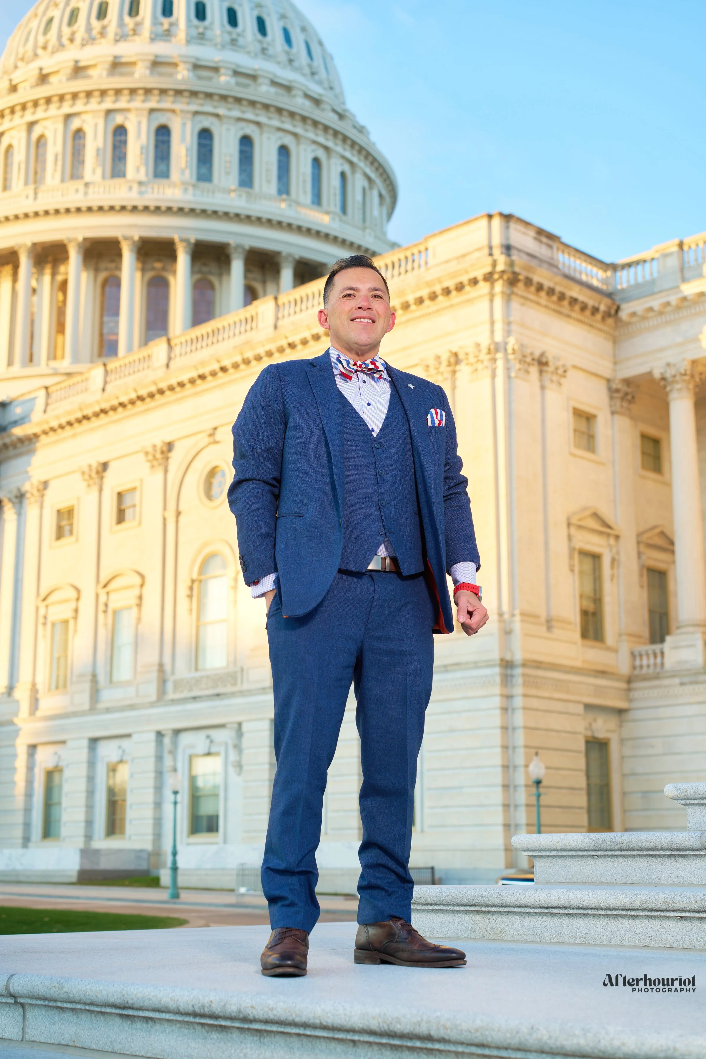 A man in a blue suit with a red, white, and blue bow tie stands outdoors in front of a large, historic government building with columns and a dome, during daylight.